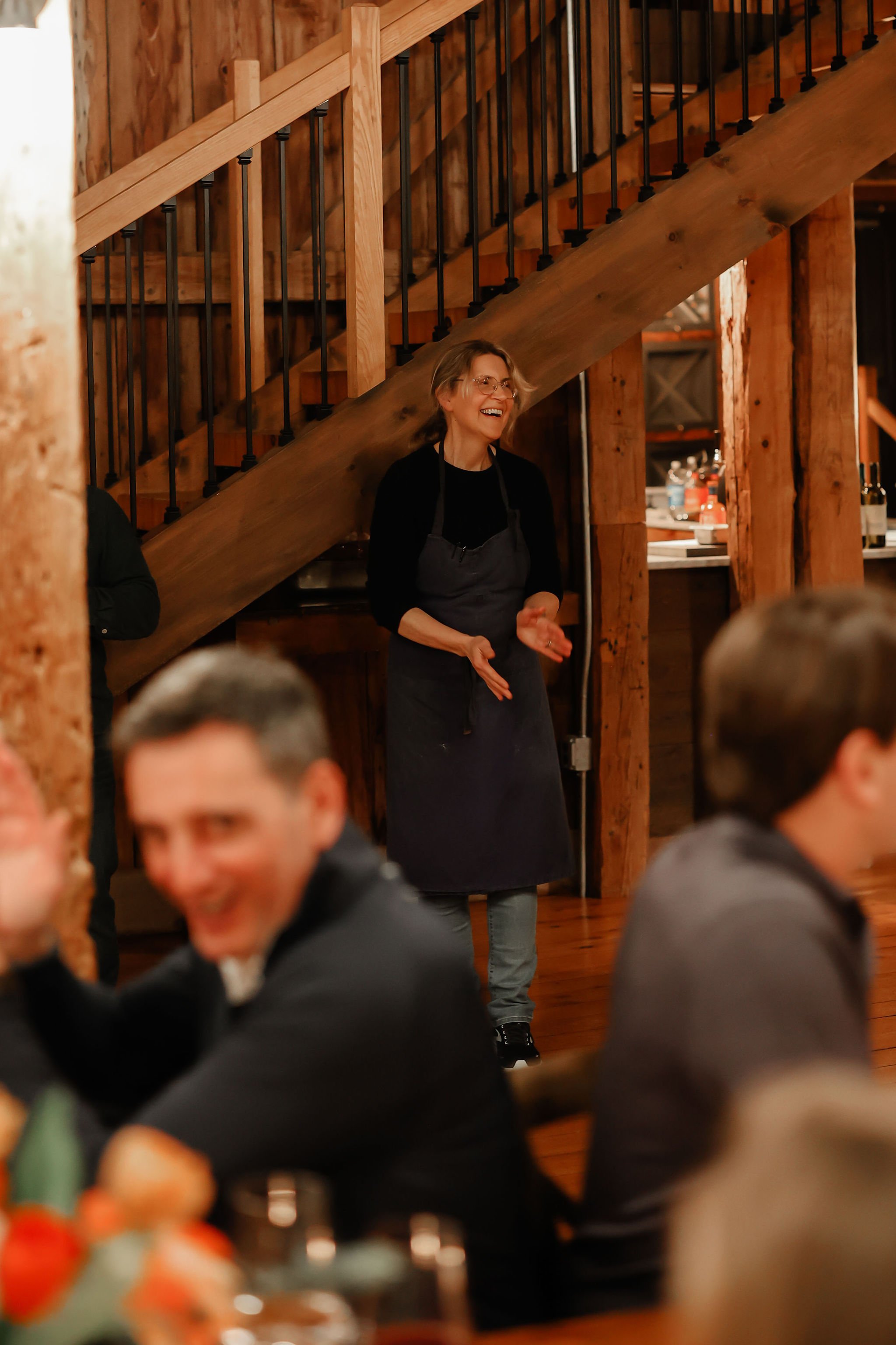 A woman standing and smiling while clapping at a restaurant or event with wooden interior and stairs in the background, surrounded by seated guests.