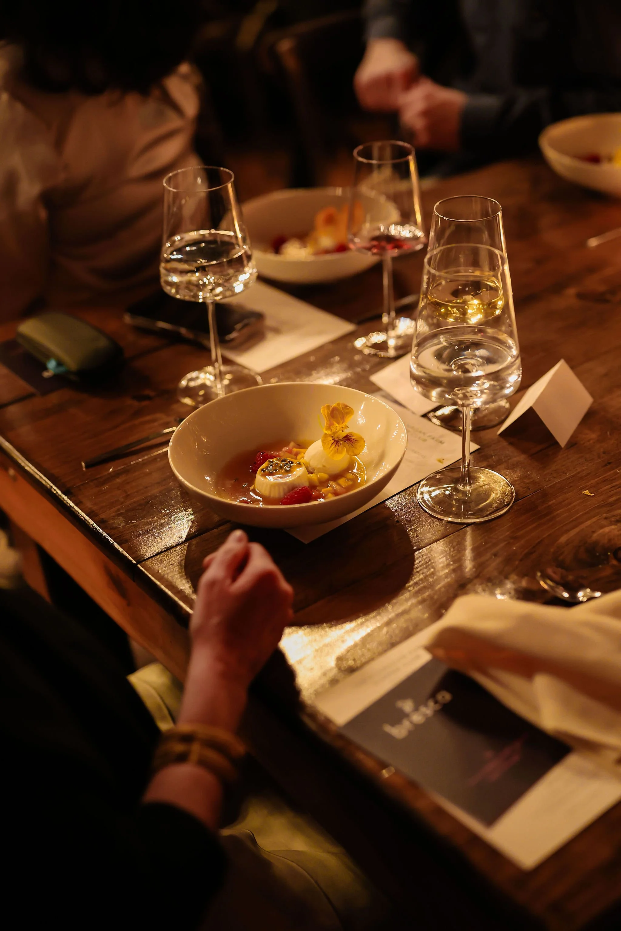 A wooden dining table set with four glasses of white wine or champagne, two bowls of dessert with fruit and edible flowers, and some utensils and napkins.