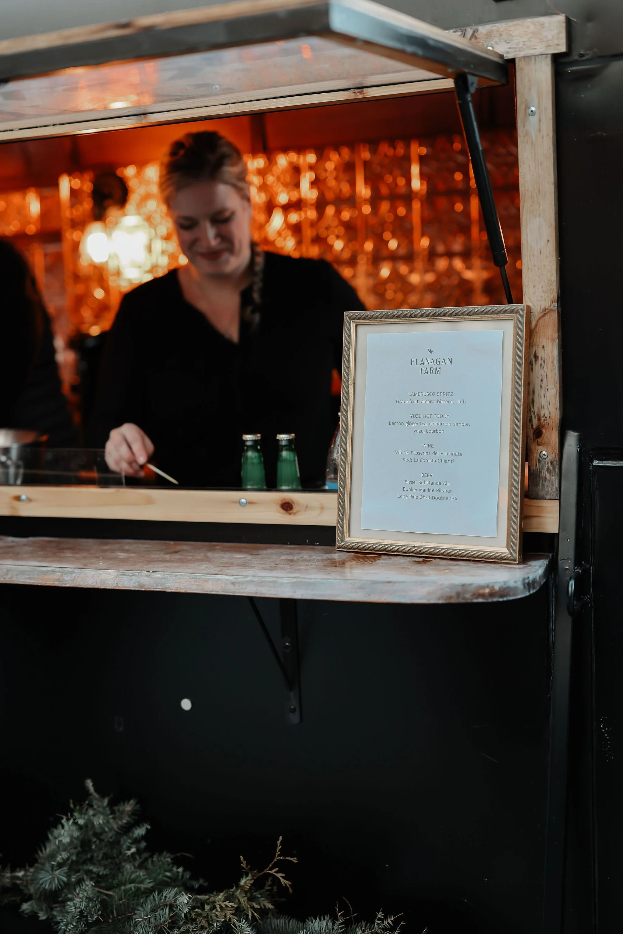 A woman working at a wooden food or drink stand with a framed menu on the counter, and illuminated decorations in the background.