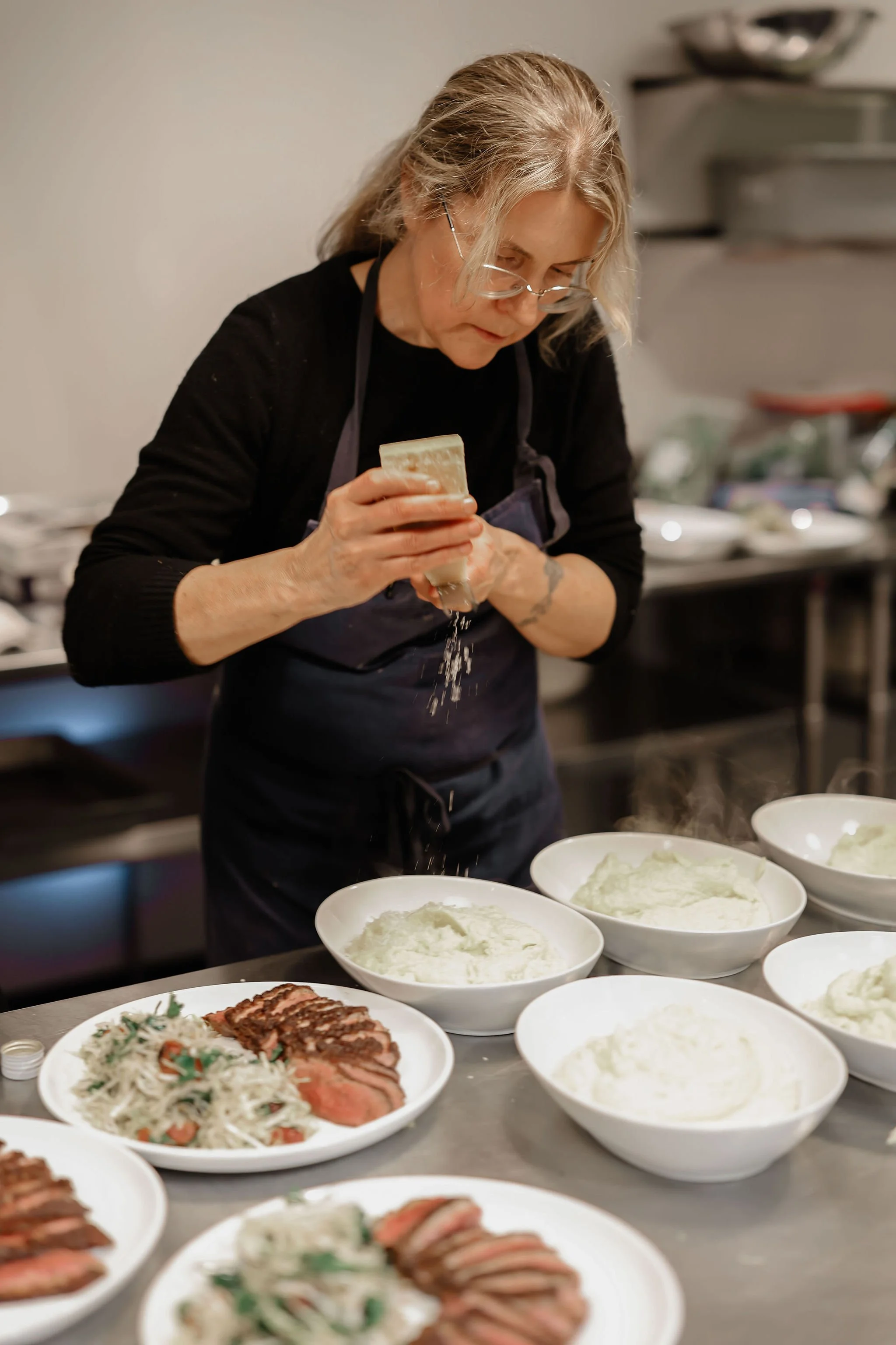 A woman in a black shirt and apron grates cheese over plates of cooked meat and mashed potatoes in a kitchen.