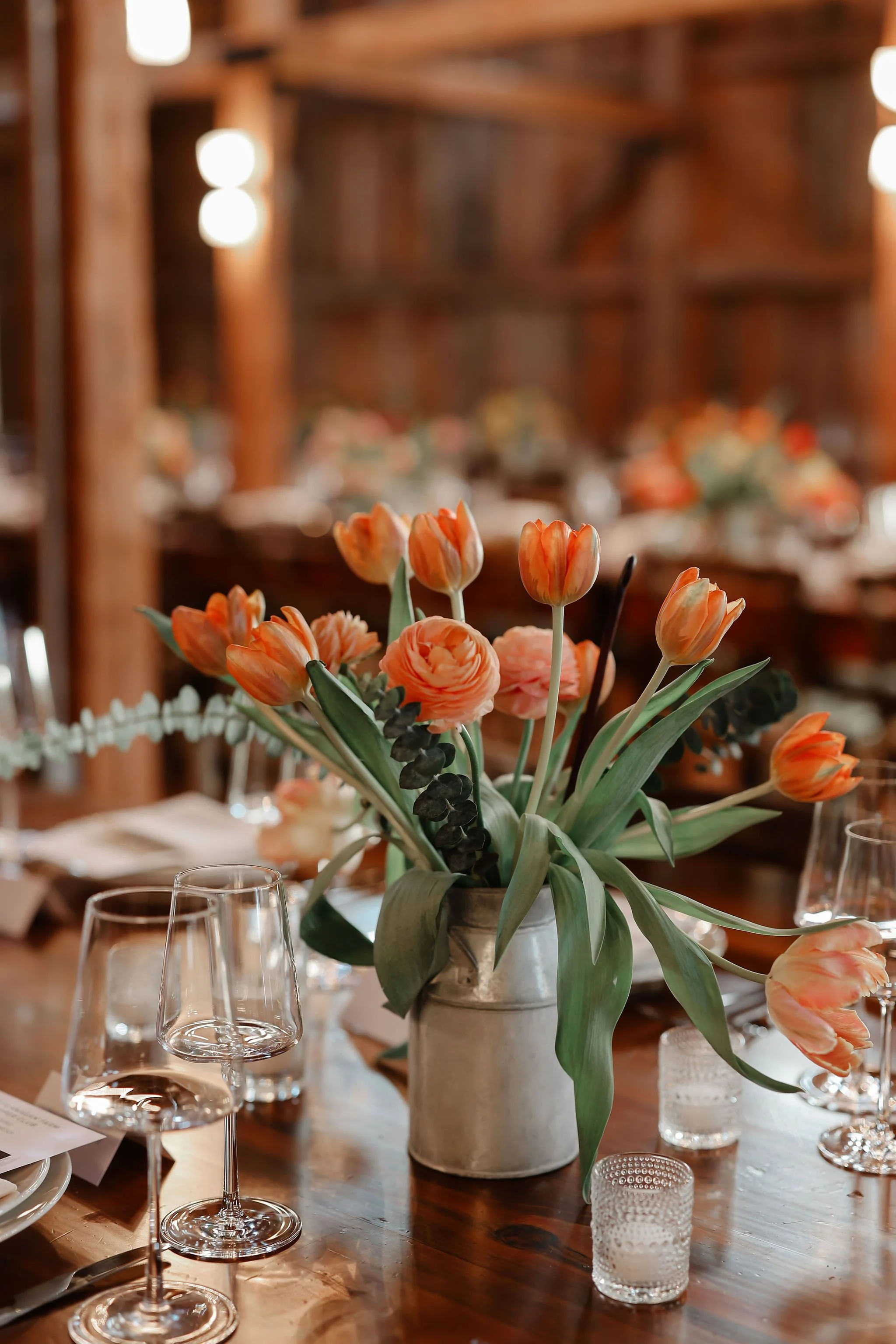 A rustic wooden table decorated with a bouquet of peach-colored tulips and ranunculus in a gray ceramic vase, surrounded by empty wine glasses and small votive candles.