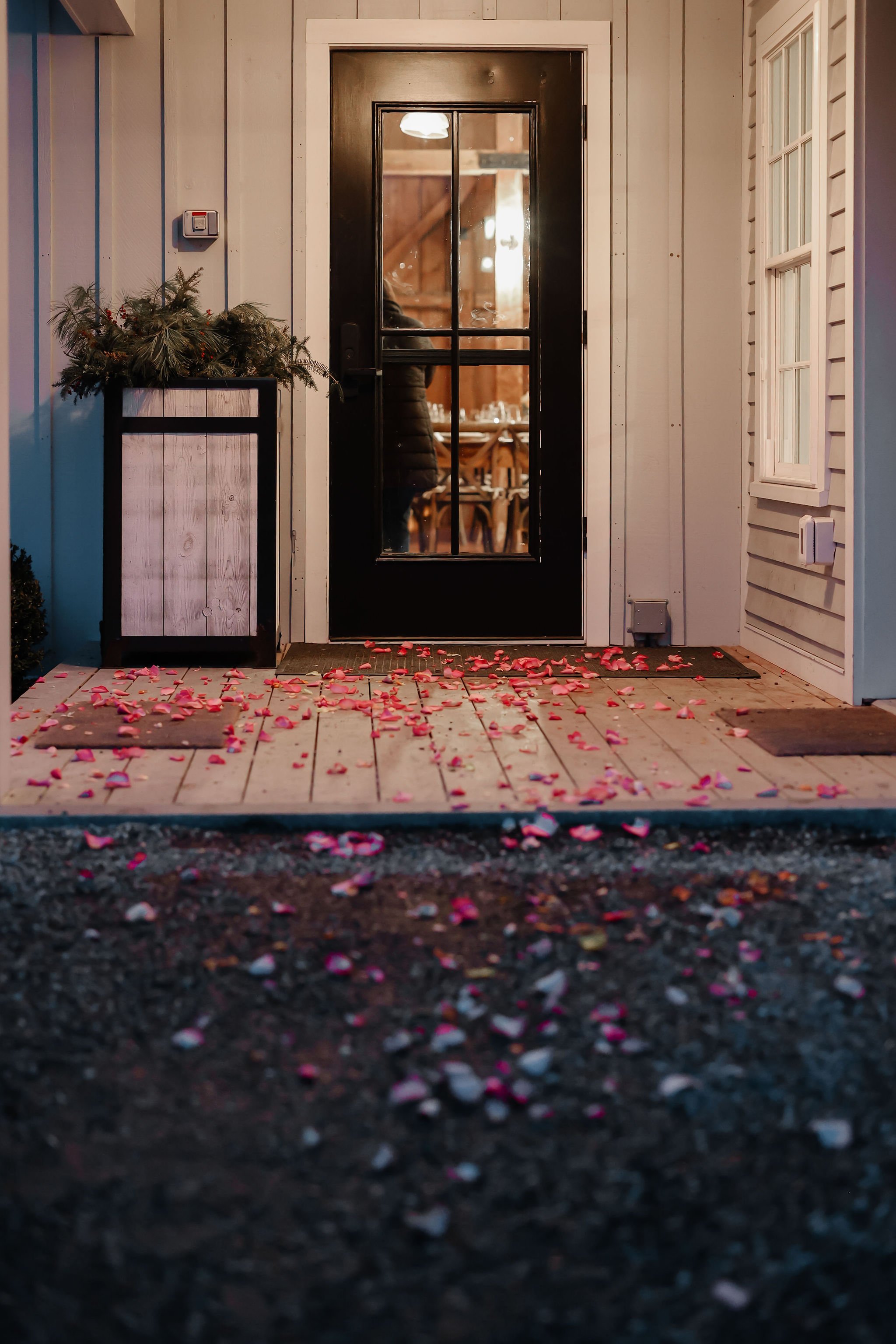 The image shows a front porch with a black door with glass panes. Rose petals are scattered on the wooden porch and the ground below. To the left, there is a potted plant with greenery and red berries, and to the right, there is a white wall with a s