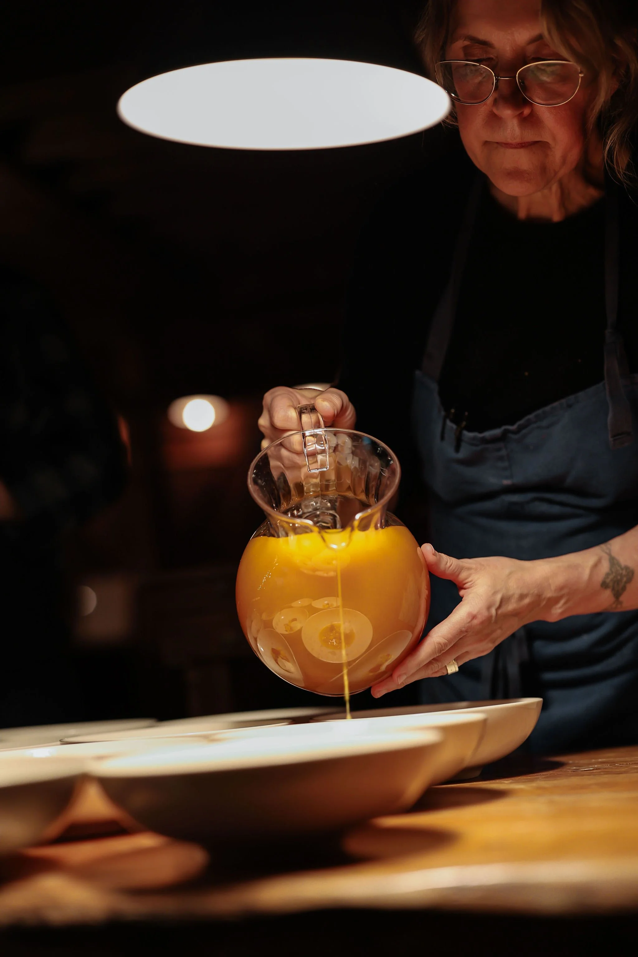 A woman with glasses and curly hair pours orange juice into a clear glass pitcher in a dimly lit room.
