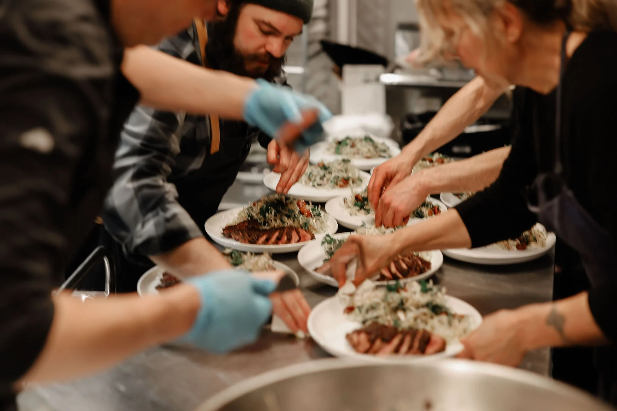 People in a kitchen preparing and plating multiple dishes of food on a stainless steel counter.
