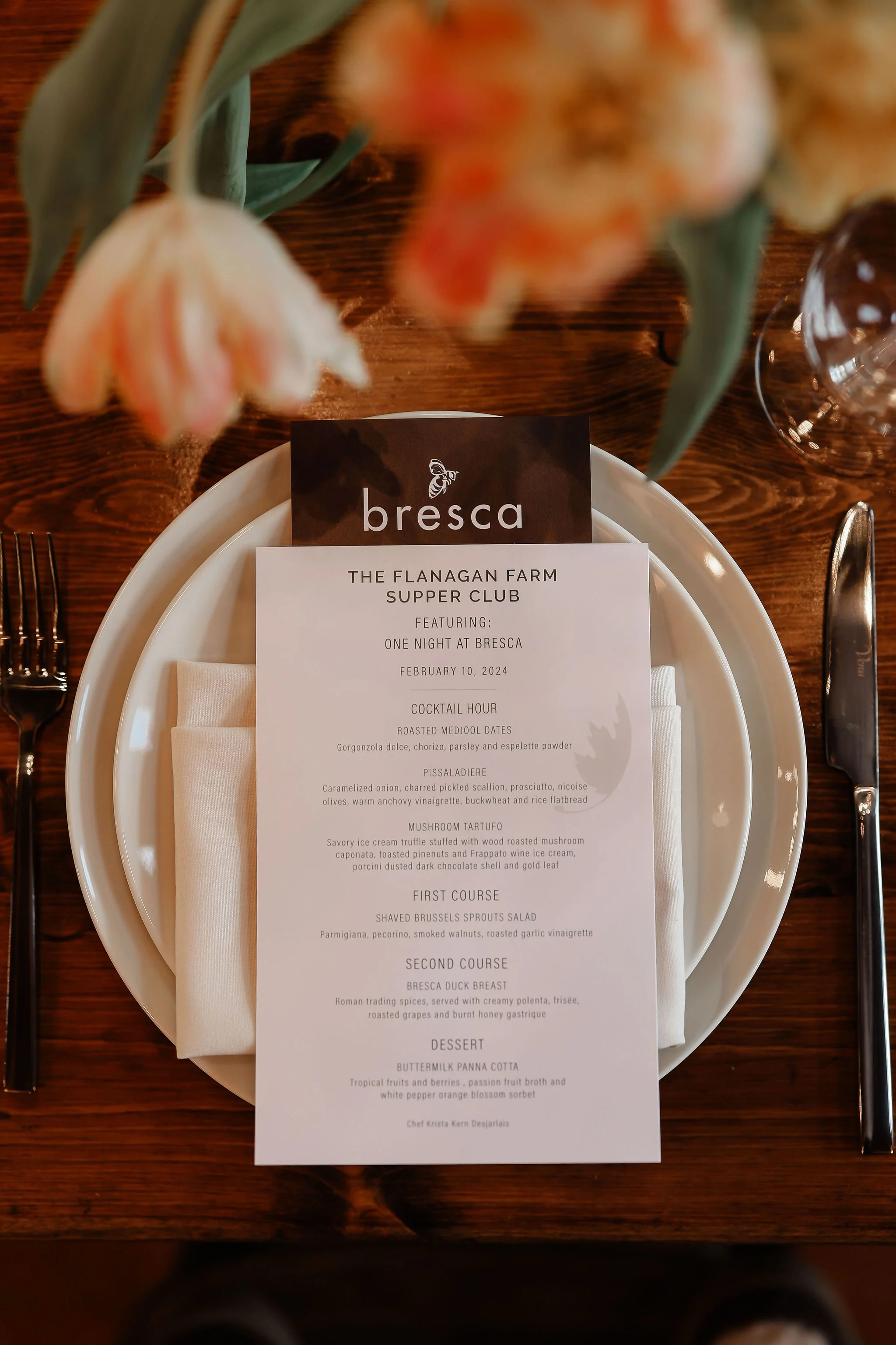 A table setting with a printed menu from Bresca, featuring a multi-course meal including appetizer, main course, and dessert, set on a wooden table with floral decorations.