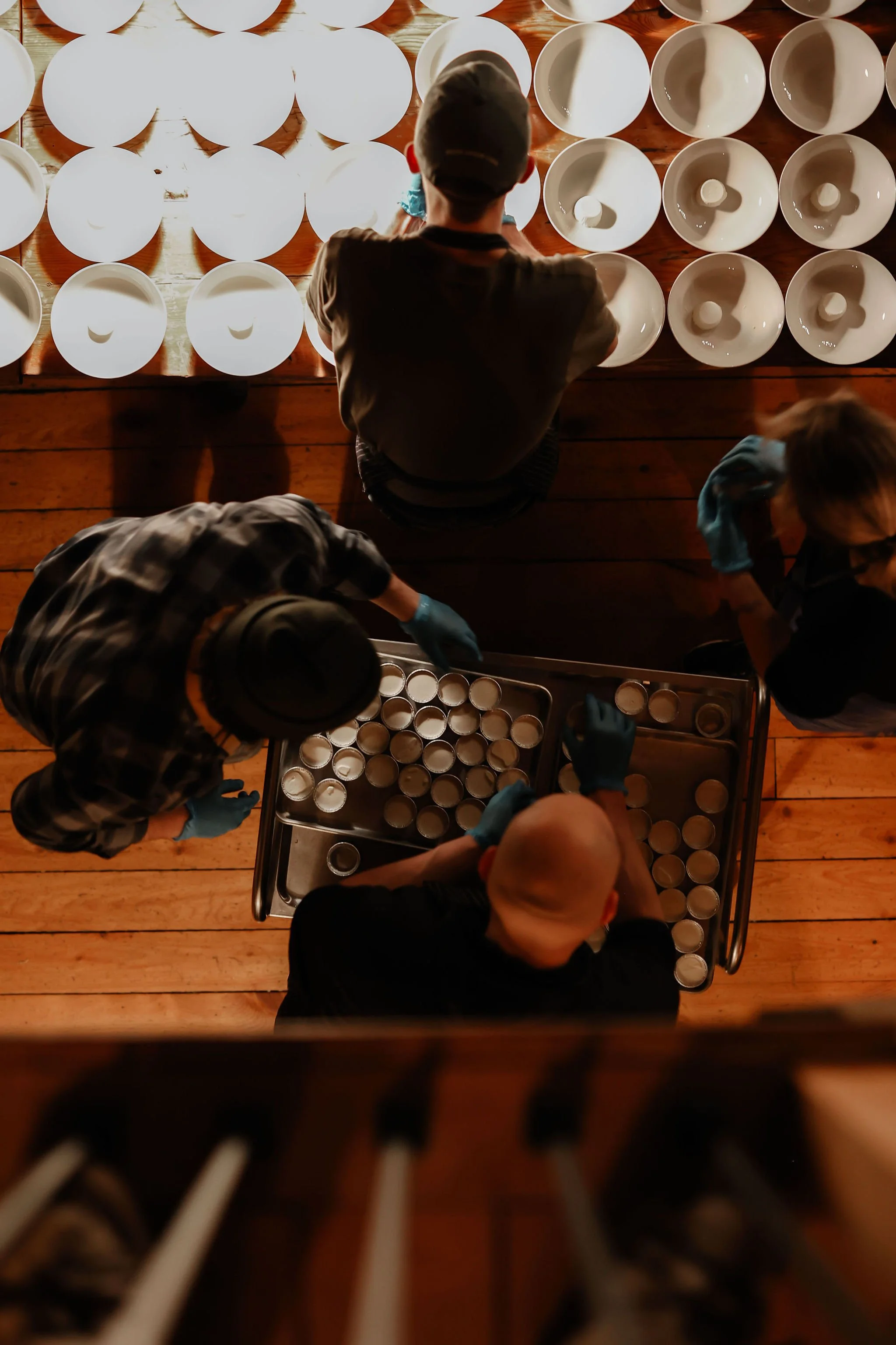 People wearing gloves working at a table filled with small white cups, with empty white plates arranged above.