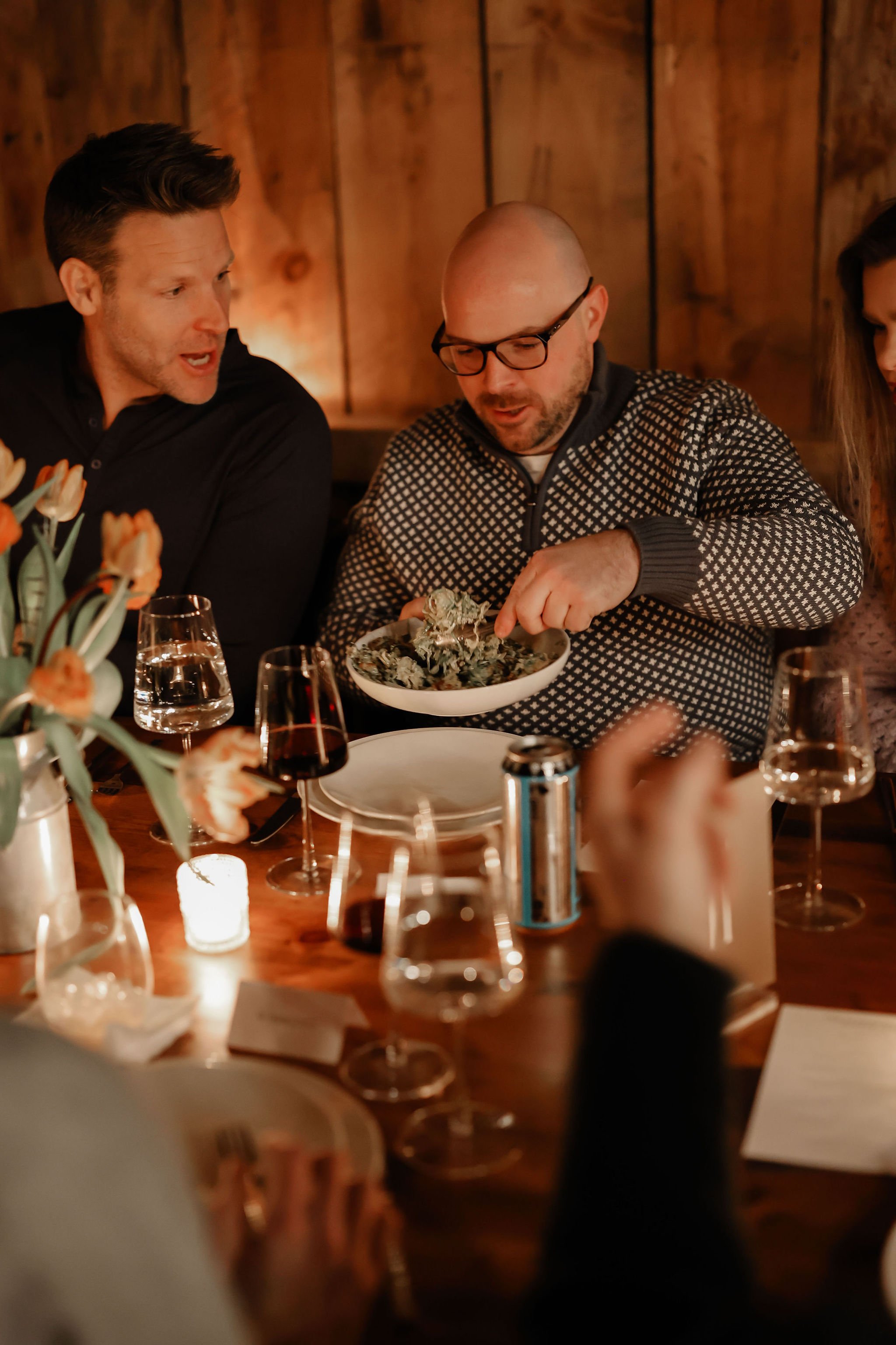 People gathered around a dinner table with wine glasses, flowers, and a candle, engaging in conversation and sharing food.