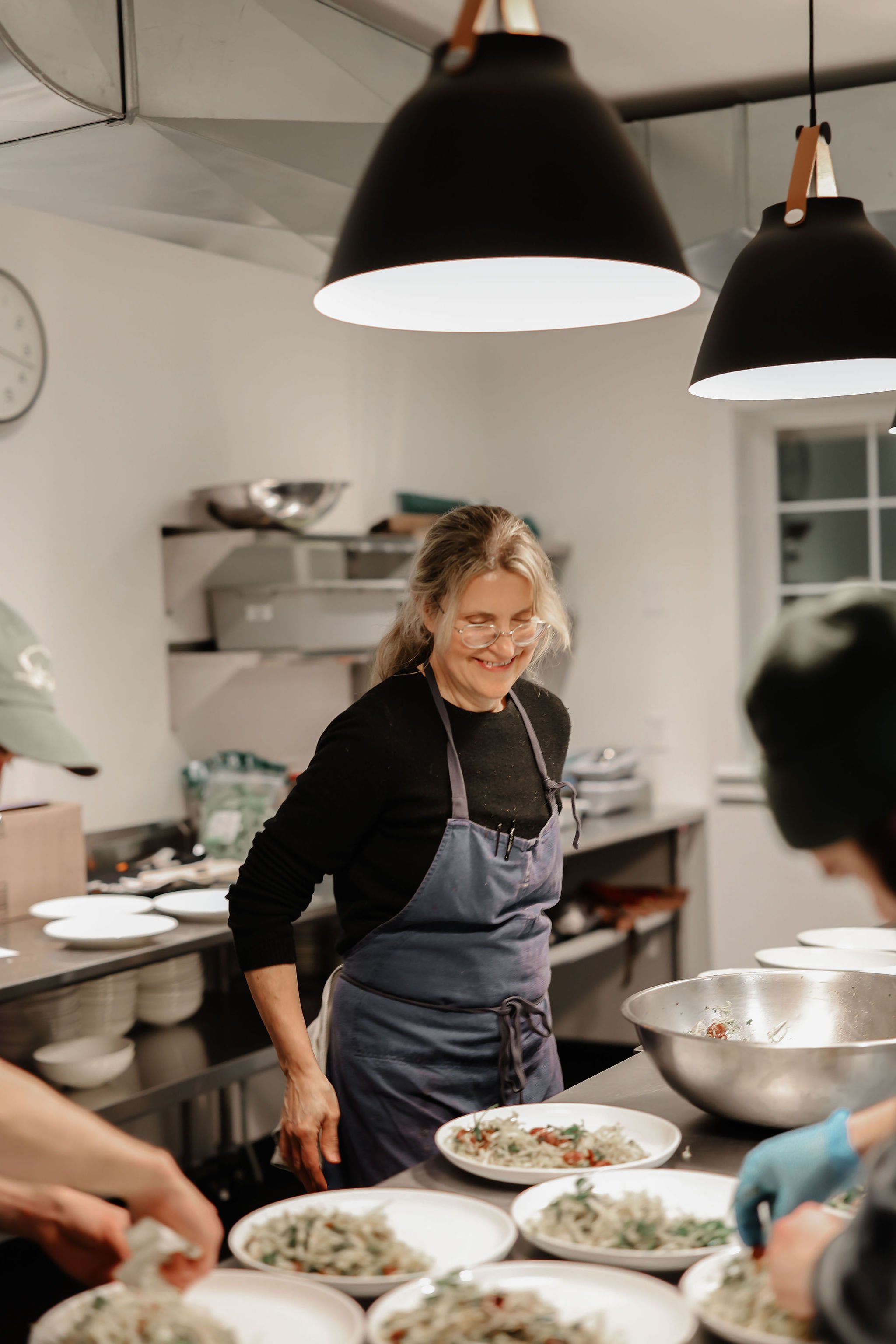 A woman with glasses and long hair, wearing a dark shirt and apron, smiling while preparing dishes in a kitchen with plates of food on the counter.