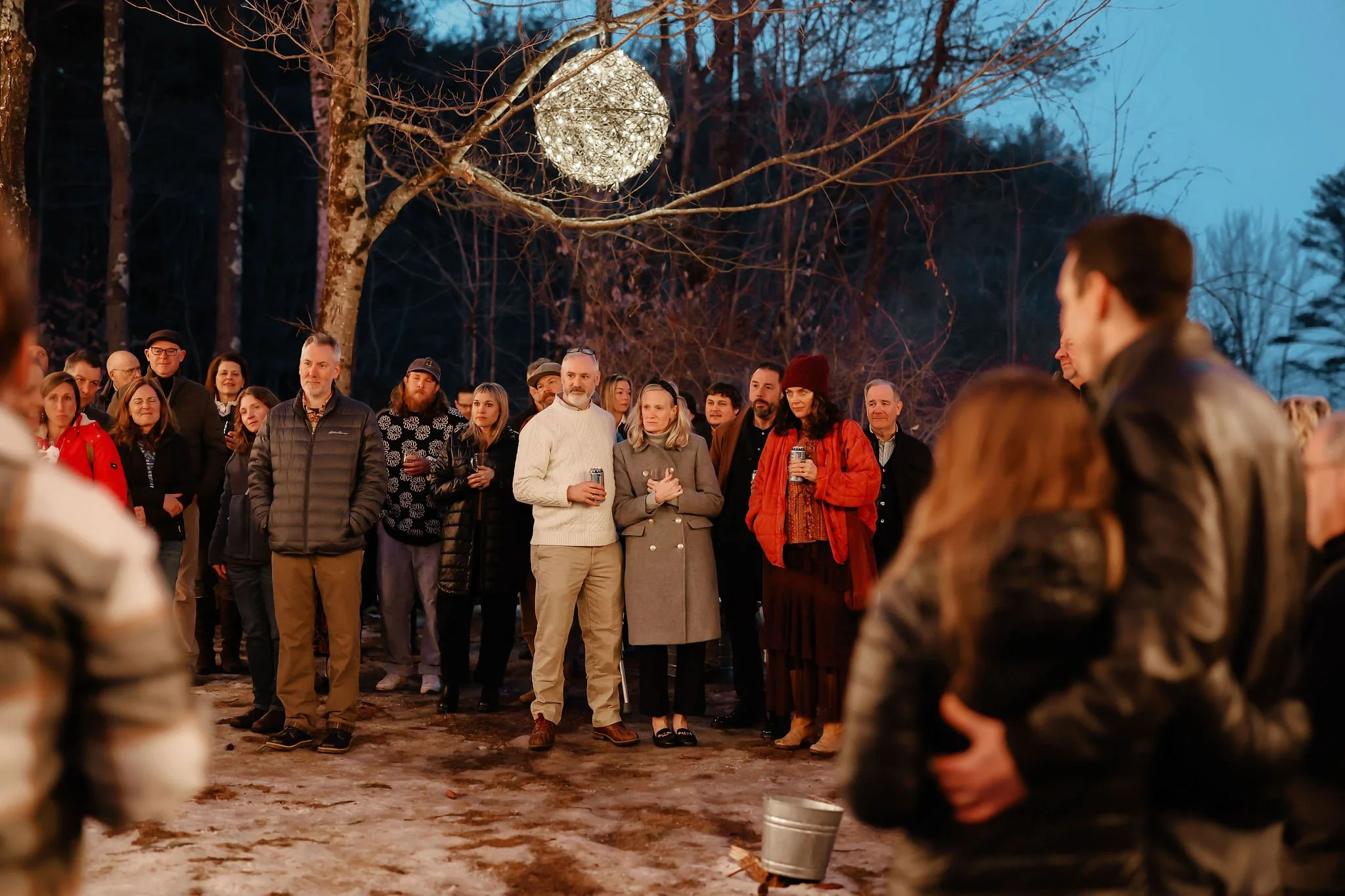 Group of people gathered outdoors at dusk, standing on snow-covered ground, with a lit decorative ball hanging from a tree above them.