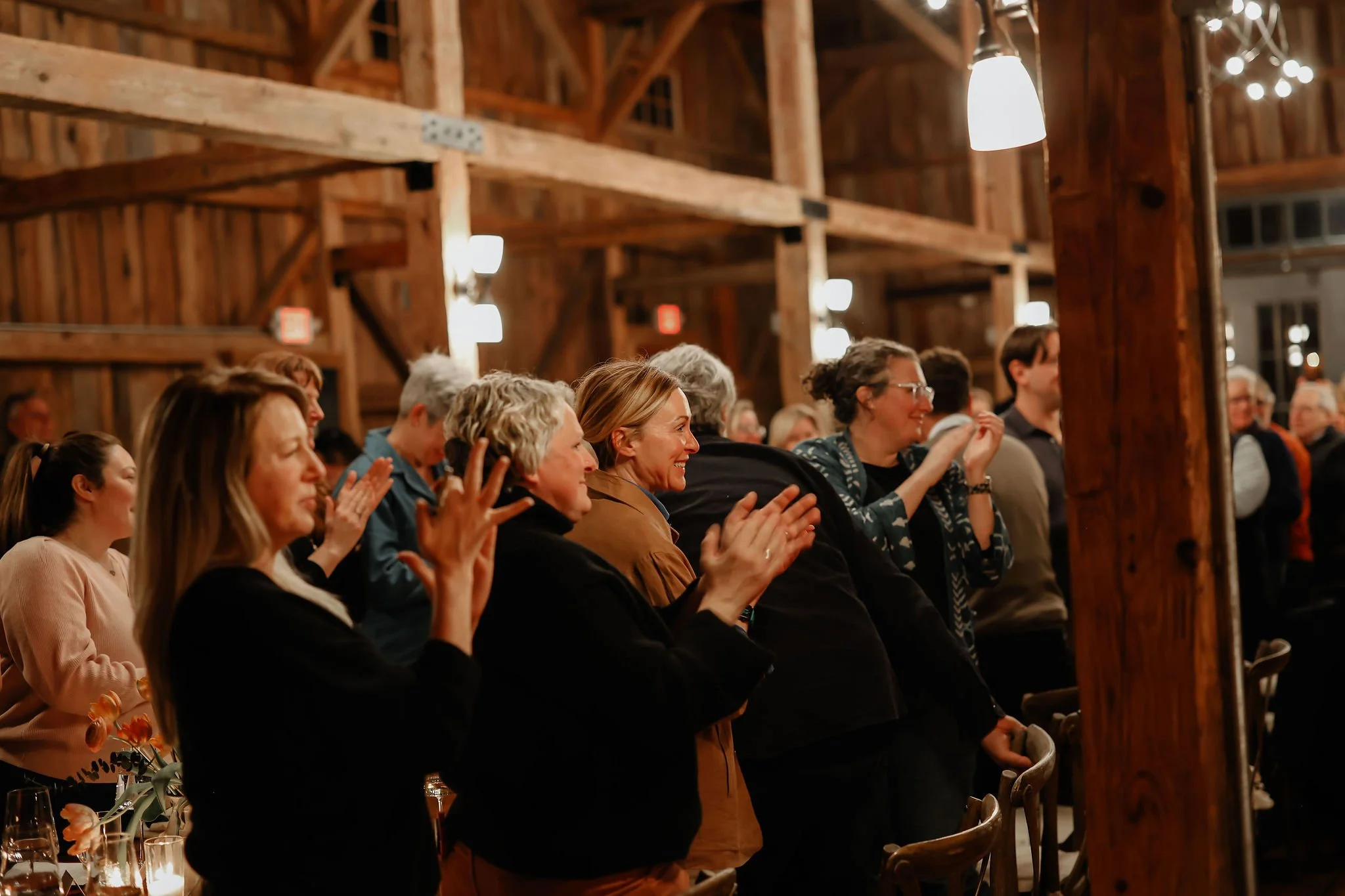 Group of people standing and clapping at an indoor event in a wooden rustic venue, with warm lighting and a joyful atmosphere.