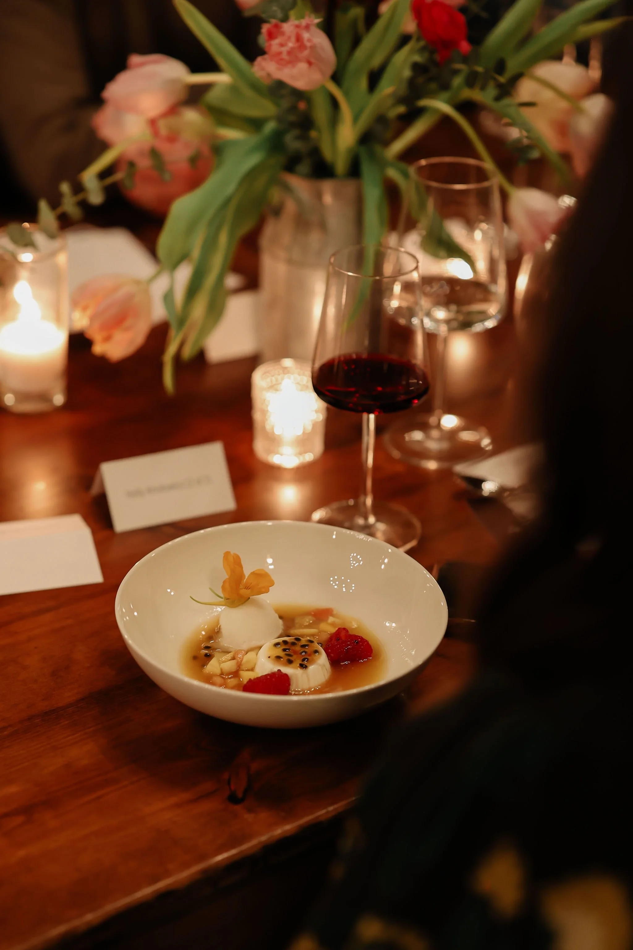 Elegant dinner setting with a small dessert in a white bowl, a glass of red wine, floral centerpiece, lit candles, and place cards on a wooden table.
