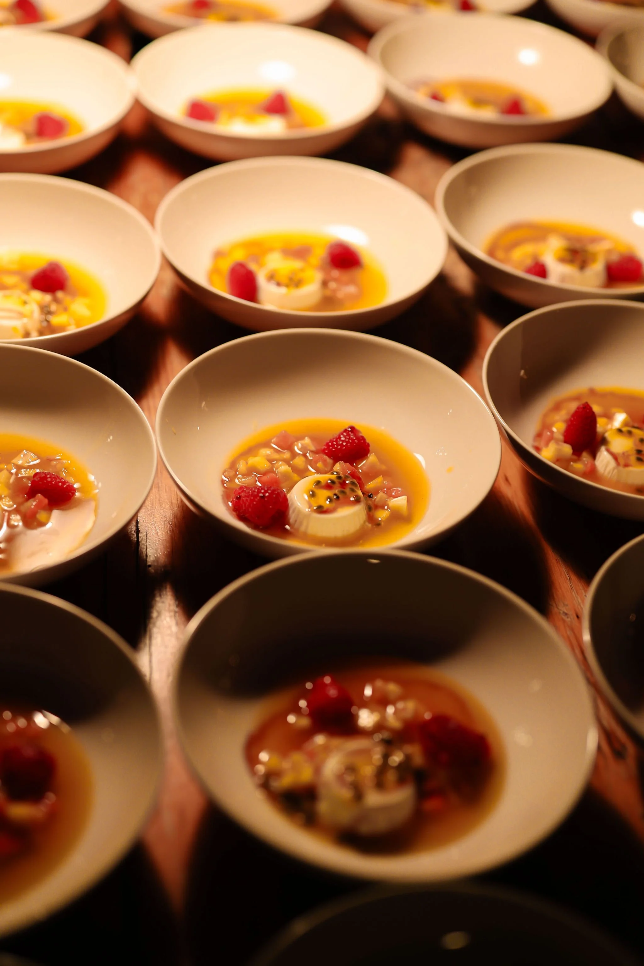 Several white bowls on a wooden surface, each containing a dessert with fruit toppings, possibly panna cotta or mousse, garnished with raspberries and other fruit pieces.