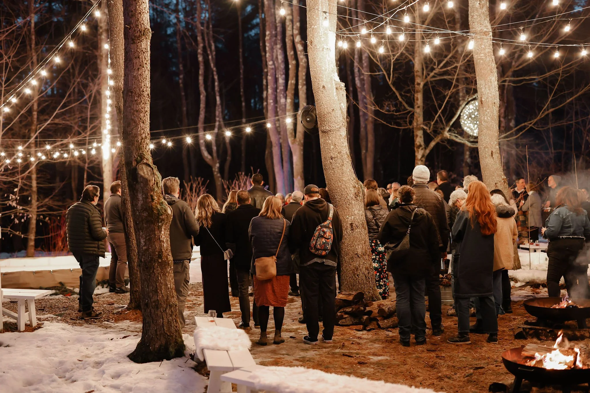 A group of people gathered outdoors in a wooded area during winter, illuminated by string lights hanging above. The ground has patches of snow, and there are several fire pits burning. The scene appears to be a social event or gathering at night.