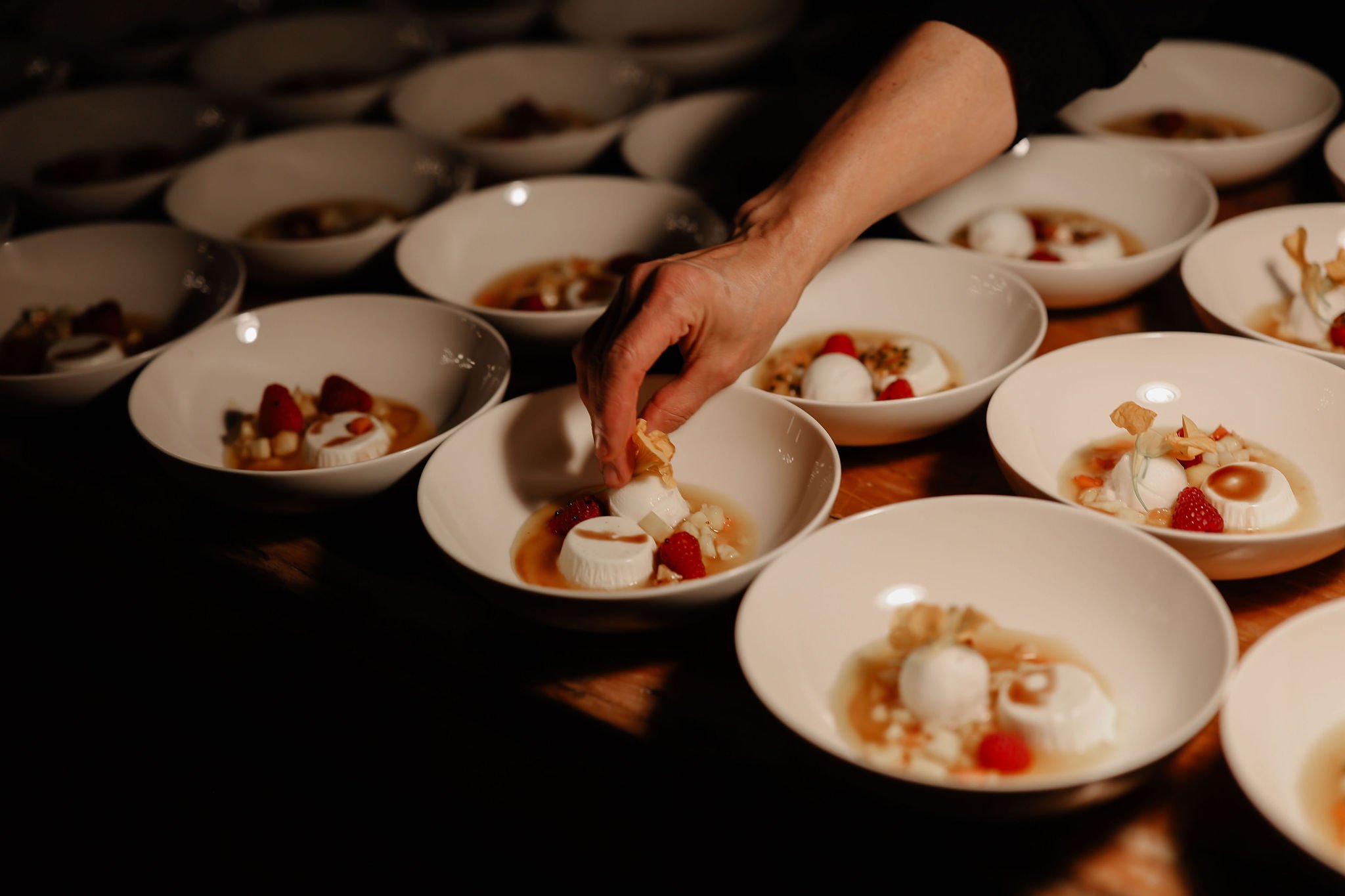 Chef garnishing plates of desserts with raspberries, white meringue, and caramel sauce in a dimly lit kitchen.