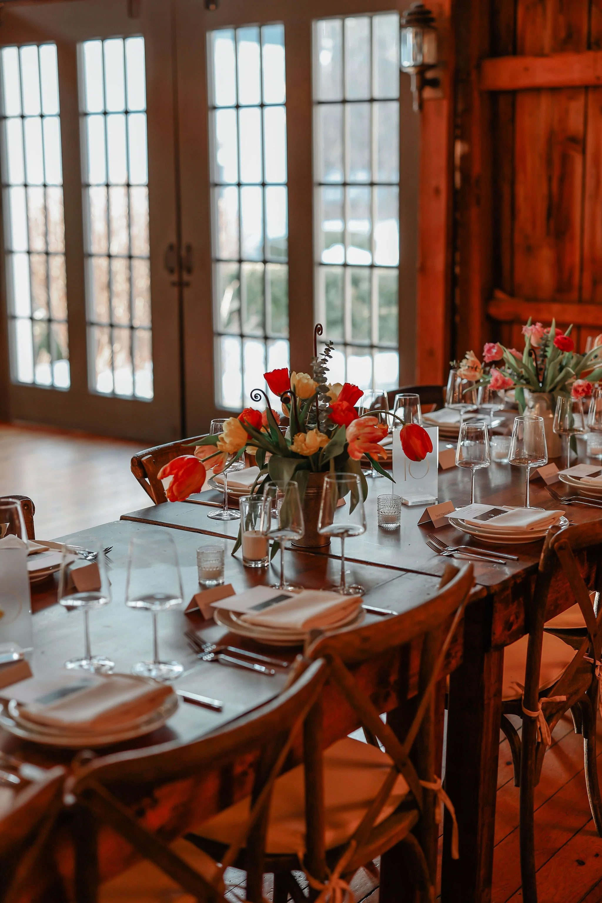 A cozy dining room setup with a long wooden table decorated with floral centerpieces, wine glasses, plates, cutlery, and napkins in front of large glass doors with a winter view outside.