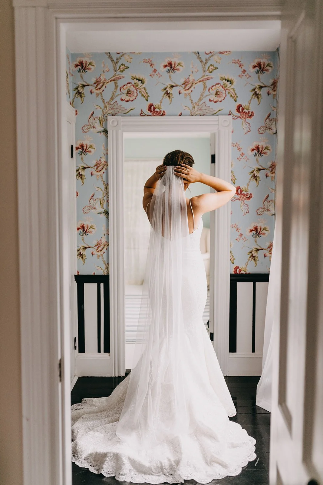 Bride in a white wedding gown and veil standing in front of a mirror, adjusting her hair in a room with floral wallpaper.