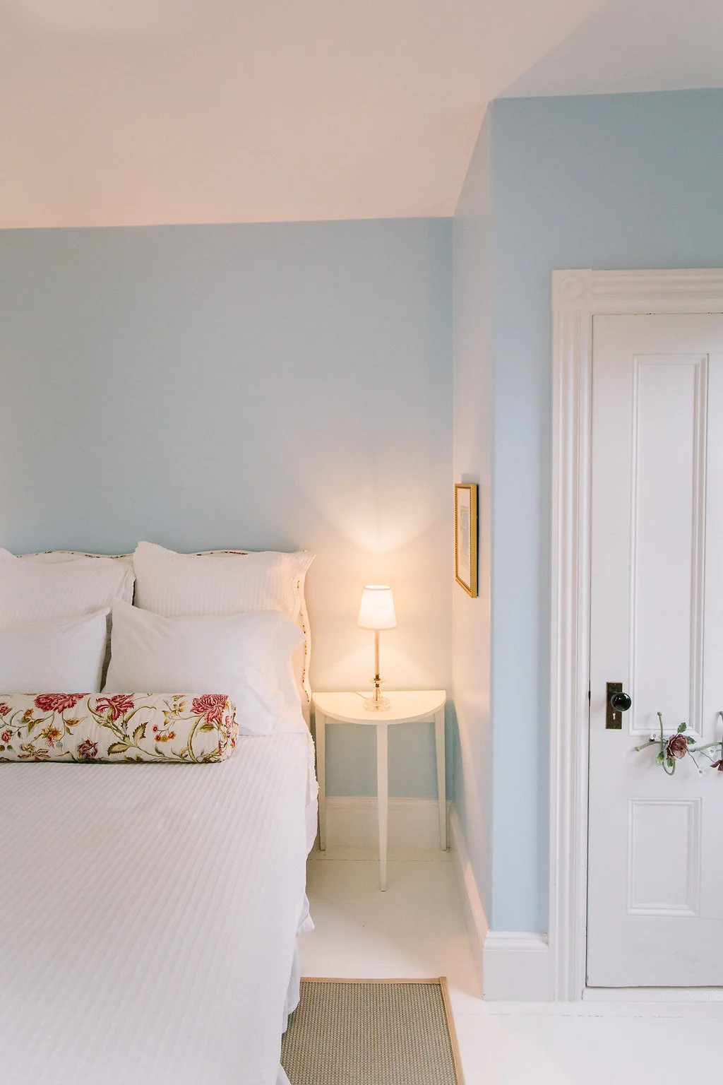 A cozy bedroom corner with a white bed, floral bolster pillow, bedside lamp, and white door with floral wreath decoration.