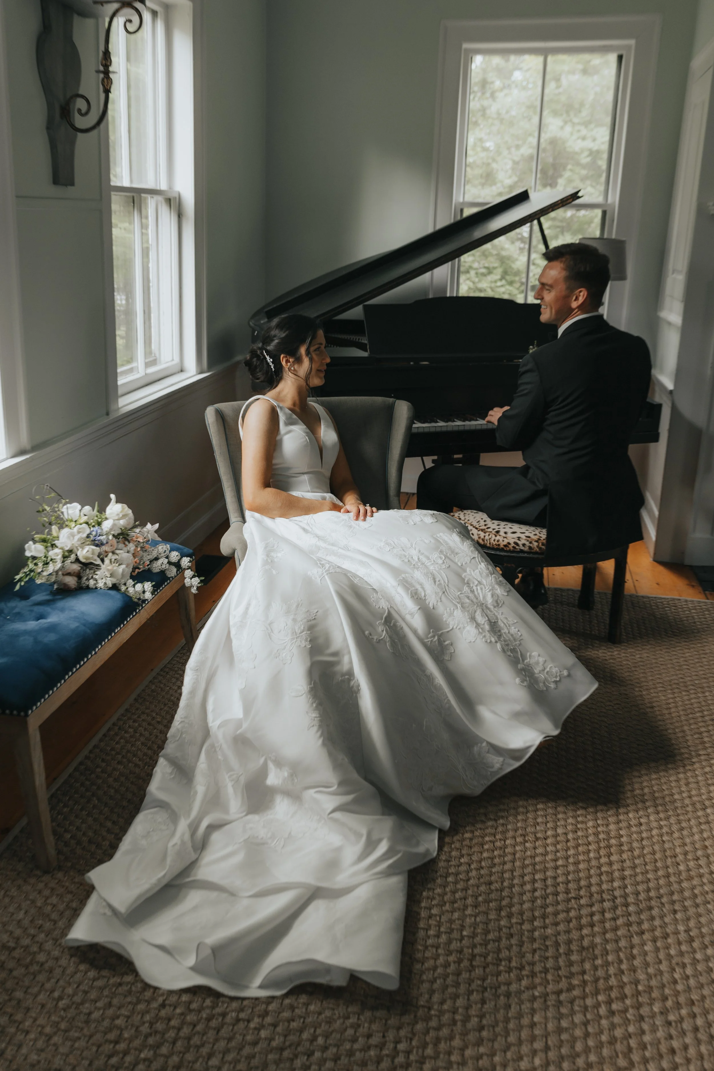 A bride in a white wedding gown sitting on a chair next to a groom in a black suit, playing the piano together in a bright room with windows and a floral bouquet on a bench.