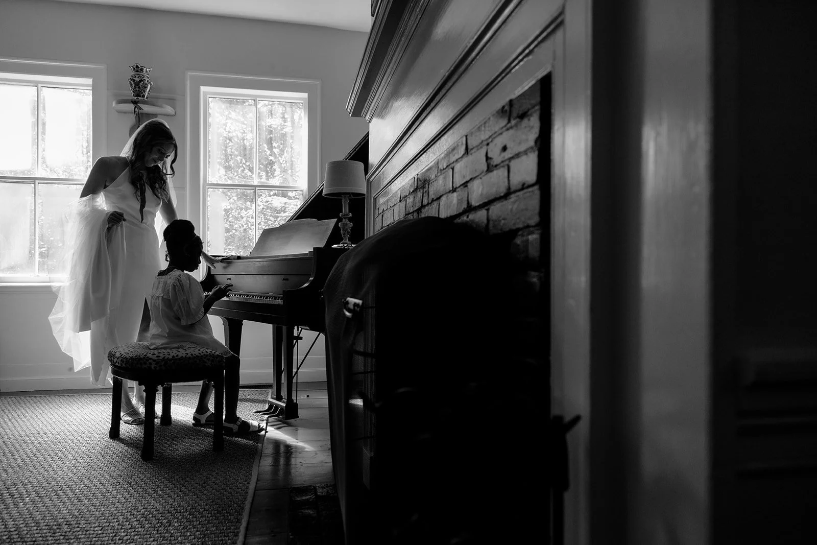 A woman in a wedding dress standing next to a young girl playing the piano in a room with windows and a brick wall.