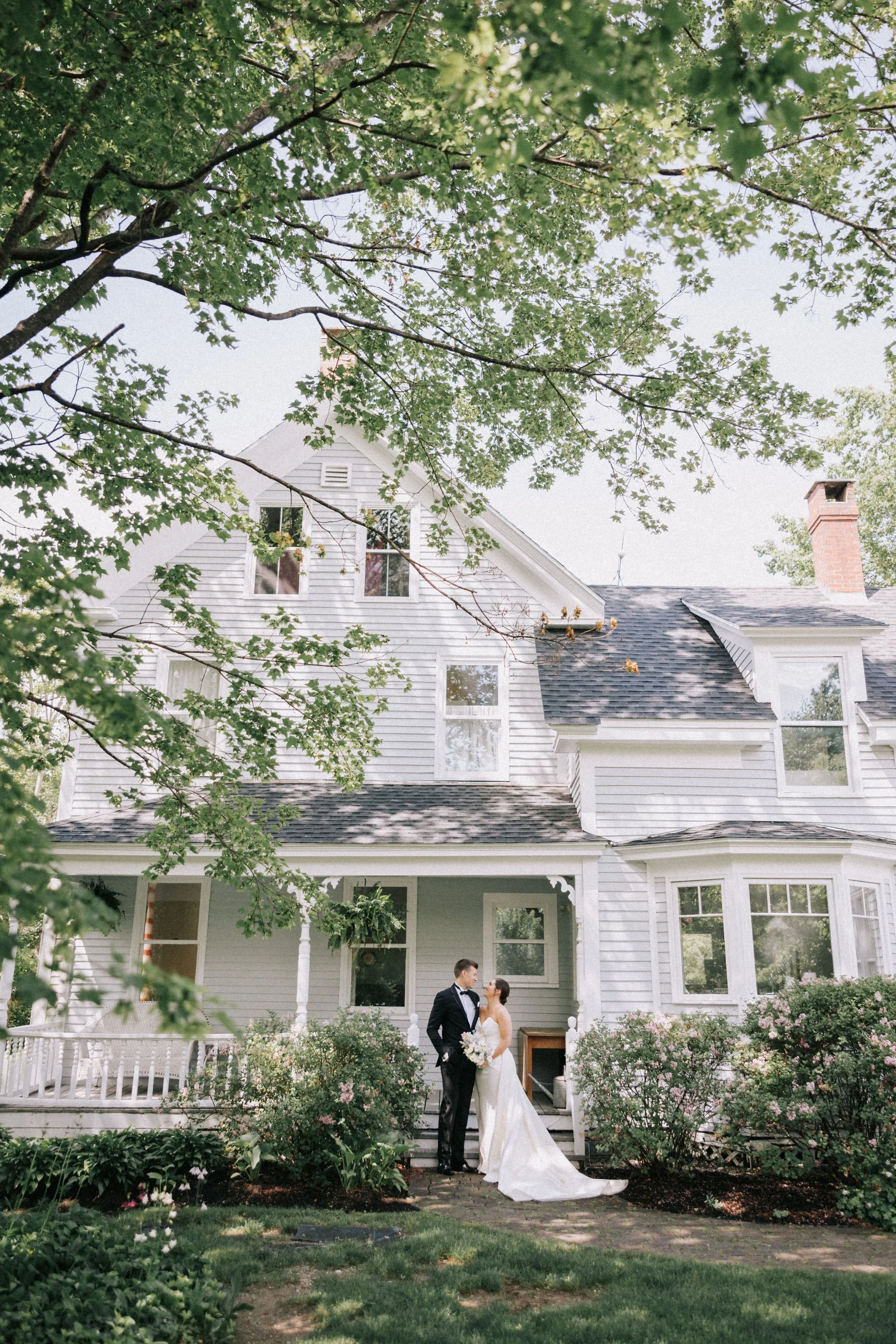 A bride and groom standing on front steps of a large white house with lush greenery and trees, enjoying a moment together on their wedding day.