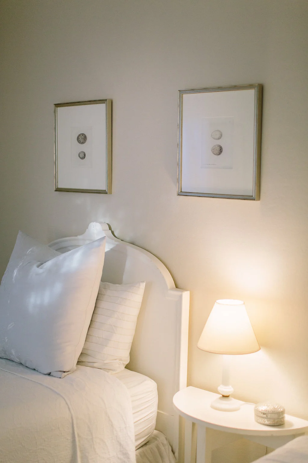 A bedroom with a white headboard, white bedding, and pillows. Two framed pictures of seashells are hung on the beige wall above the bed. A small white bedside table with a lamp and silver decorative object is positioned to the right of the bed.