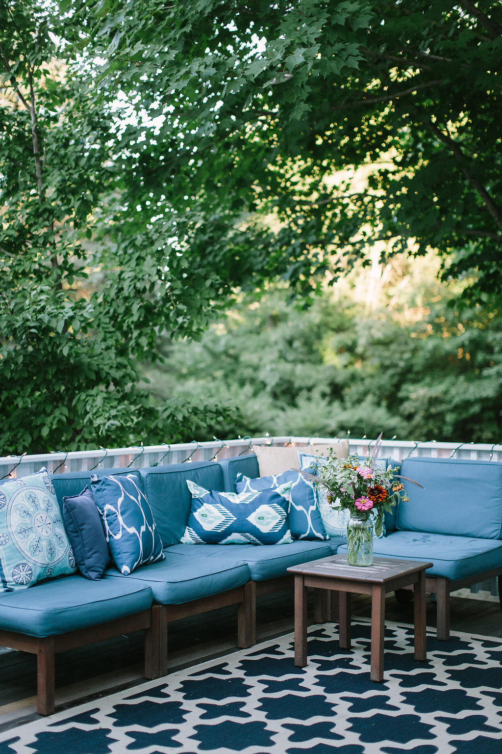 Outdoor patio with blue cushioned sectional sofa, patterned pillows, a wooden coffee table with a colorful flower arrangement, and a black and white patterned outdoor rug, surrounded by green trees.