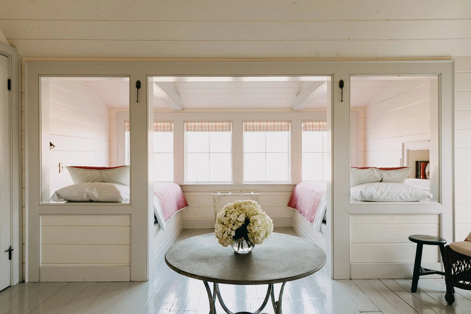 Bright room with two beds with pink blankets, flowers on a table, and windows with striped shades.