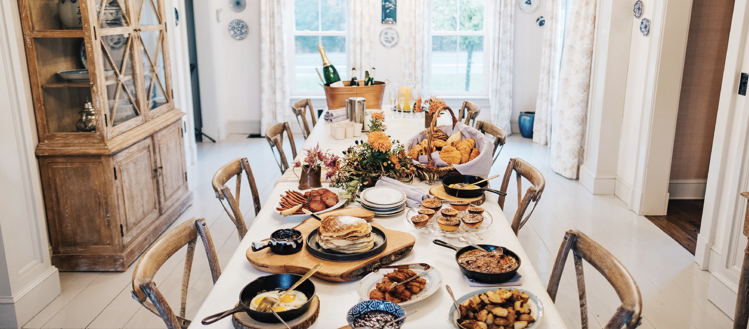 A dining table set for breakfast with various dishes, pancakes, eggs, bacon, muffins, and bread, in a bright room with large windows and floral curtains.
