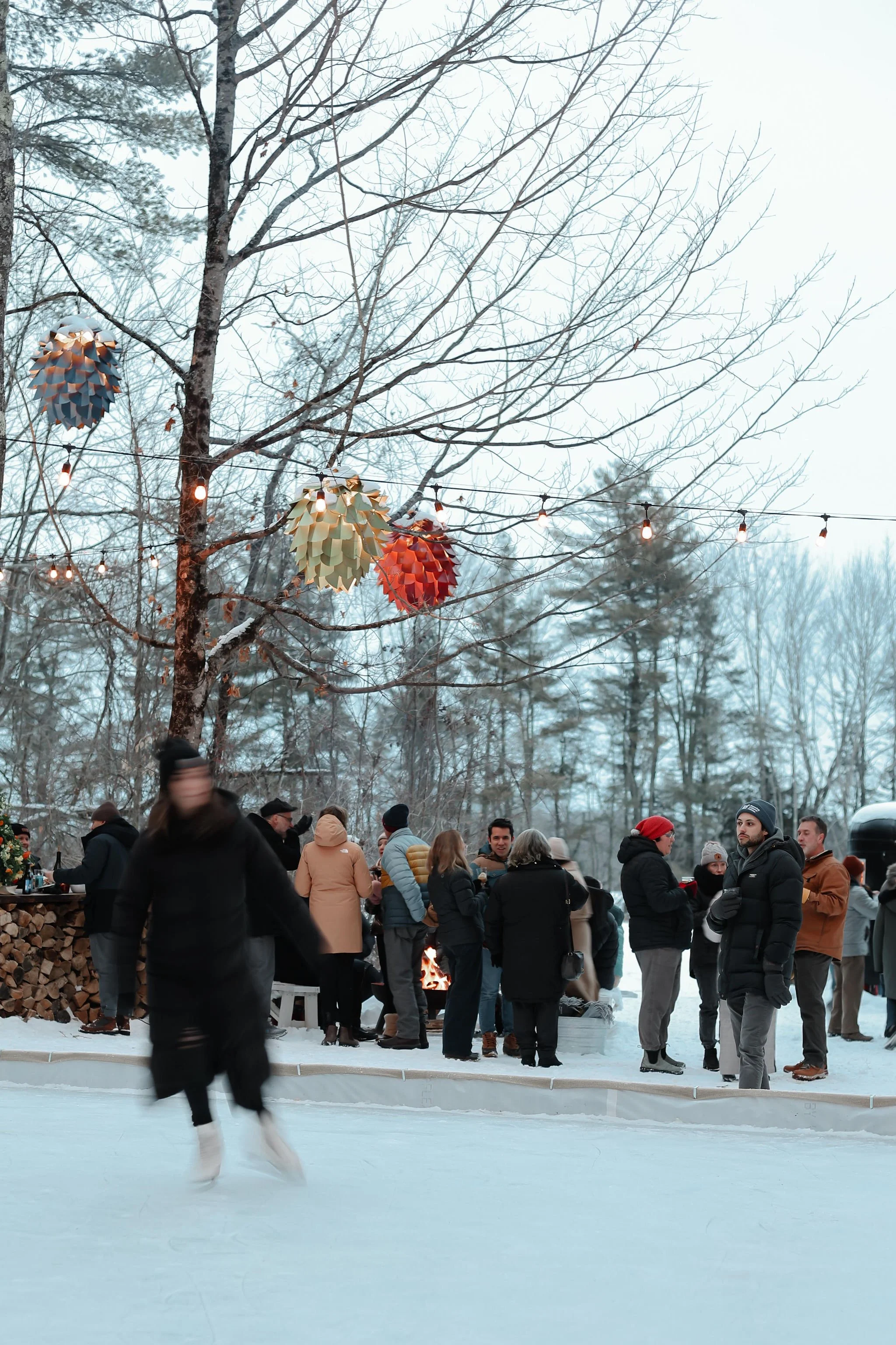 People gathered outside in winter, near a snow-covered ice rink, under a tree with colorful lanterns and string lights, with trees in the background.