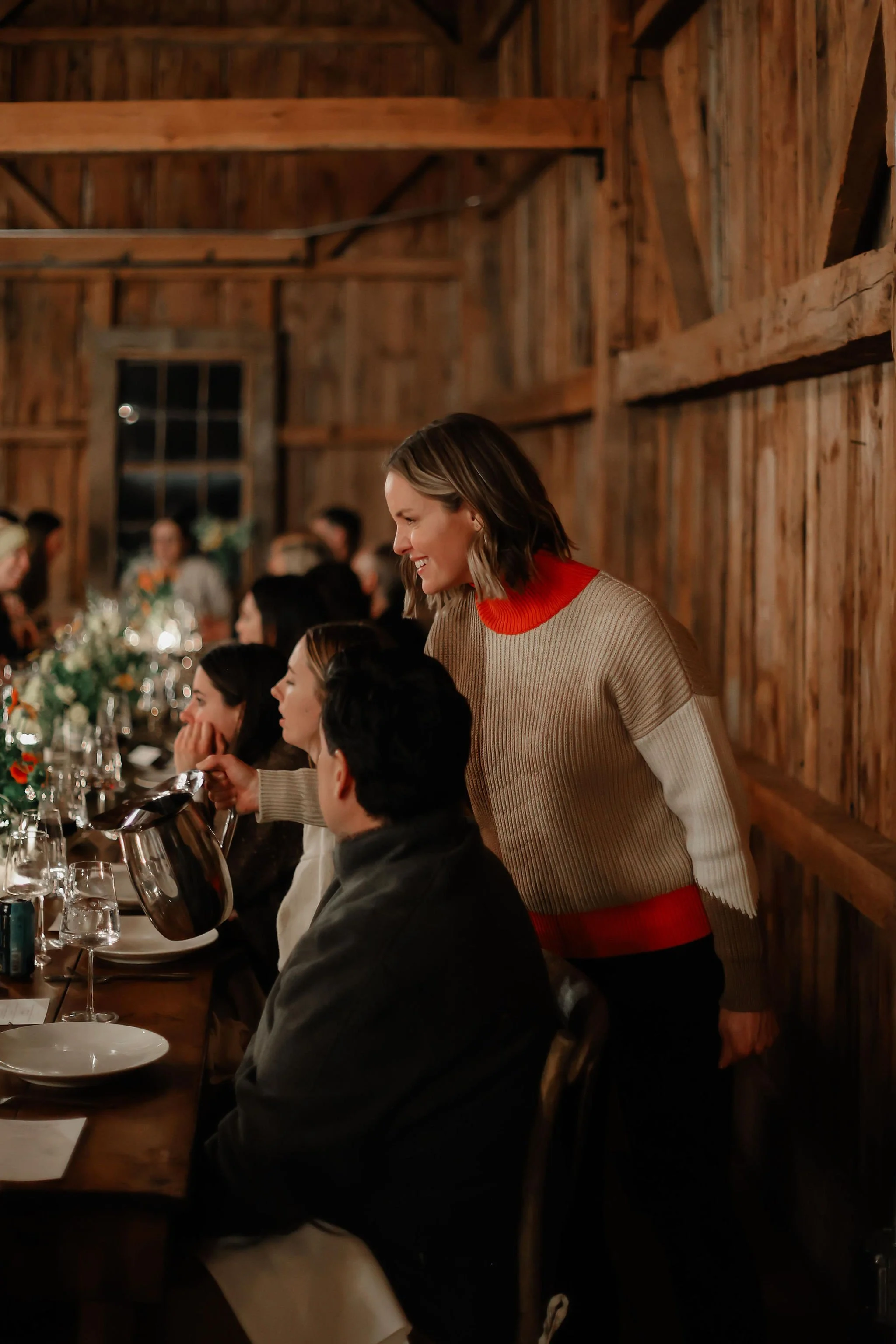 A woman in a beige and red sweater smiling and pouring wine at a rustic dinner party with several guests seated at a long wooden table decorated with flowers and candles.