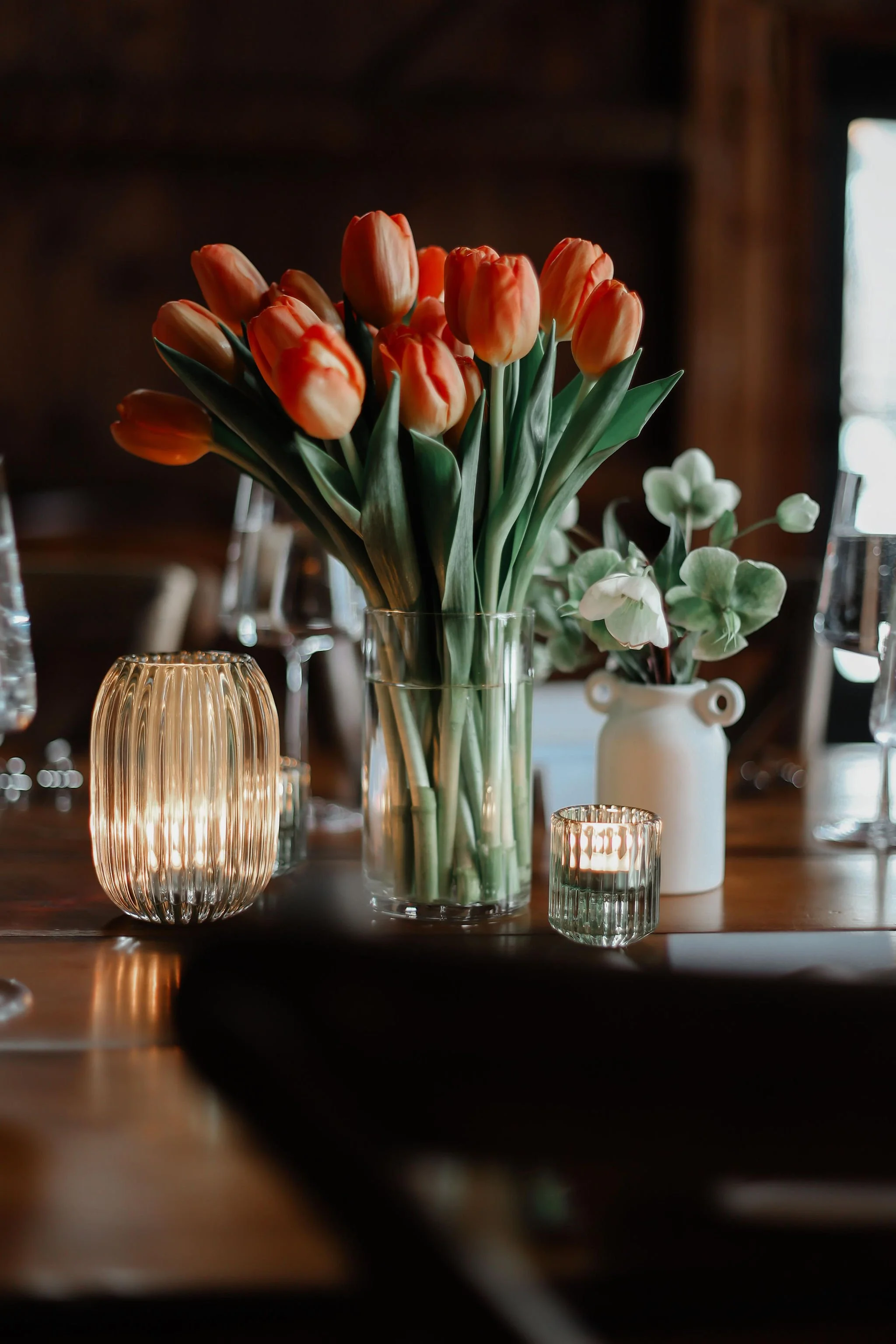A floral arrangement of orange tulips and green leaves in a clear glass vase, surrounded by small candles and a white vase with green foliage on a wooden table.
