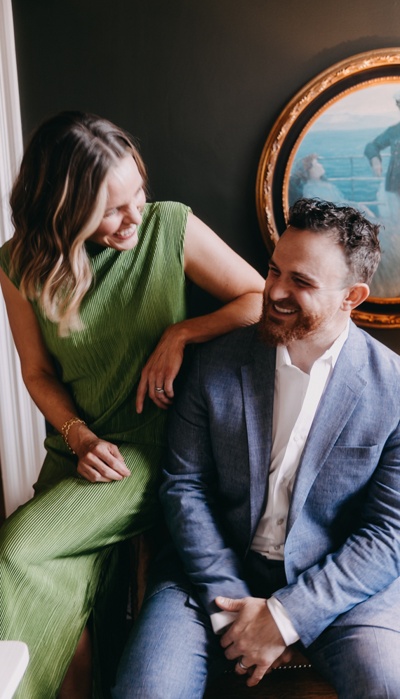 A woman and a man sharing a joyful moment indoors. The woman is wearing a green dress and leaning on the man's shoulder, both are smiling.