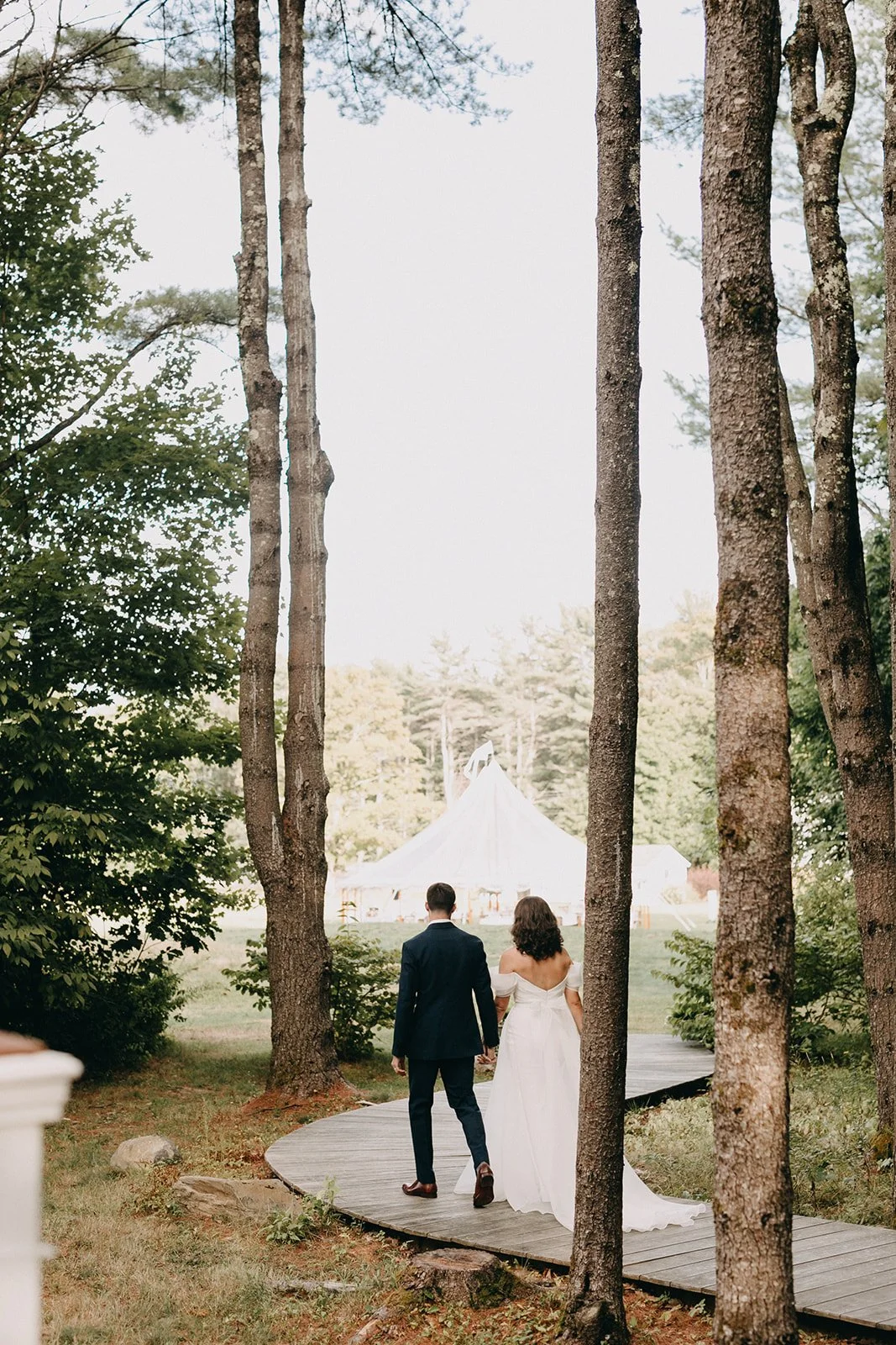 A bride and groom walking hand in hand on a wooden pathway through trees towards a white tent at an outdoor wedding venue.