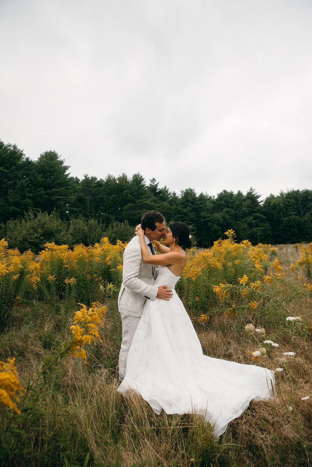 A bride and groom embrace in a field of yellow flowers on a cloudy day, with trees in the background.