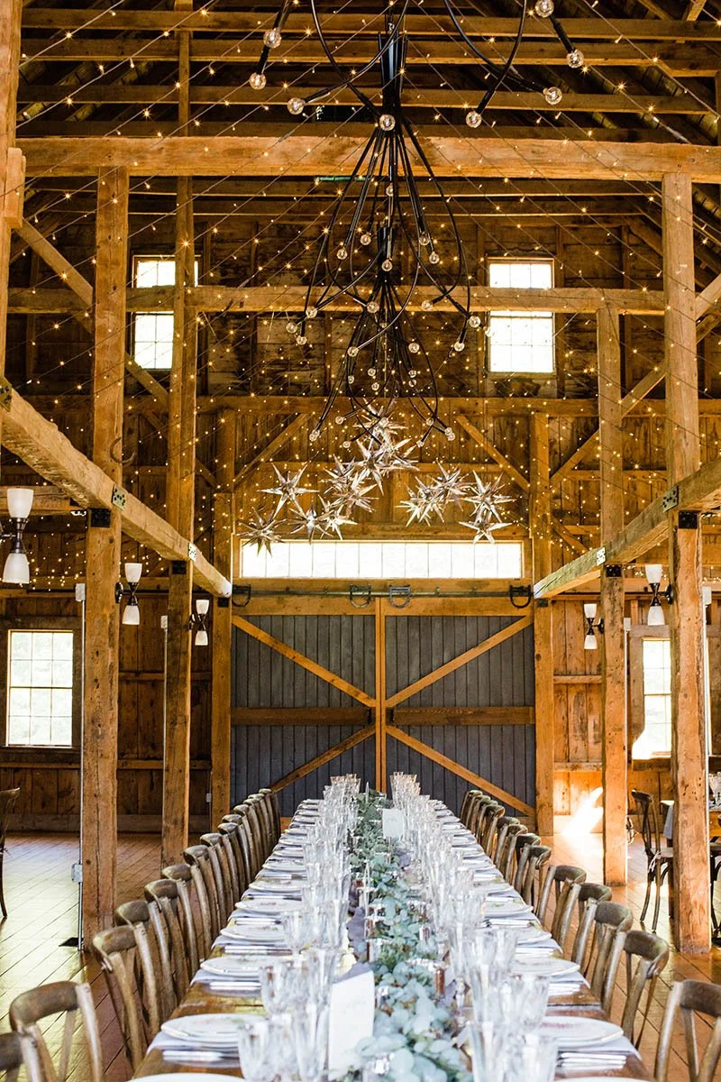 A rustic barn interior decorated for a special event with a long dining table set with glassware and napkins, a hanging star-shaped chandelier, twinkling string lights, and a large modern black chandelier with exposed bulbs.