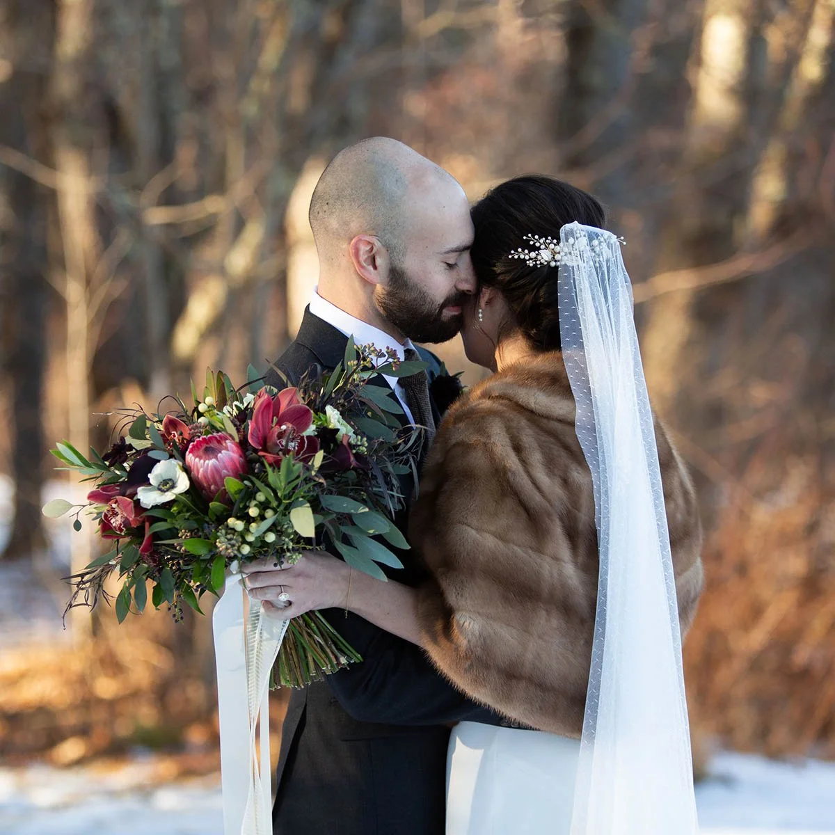 A bride and groom sharing a close moment outdoors during winter, with the bride holding a large bouquet of red, white, and green flowers. The bride wears a wedding dress, faux fur stole, and veil, while the groom is in a dark suit. Bare trees are in 