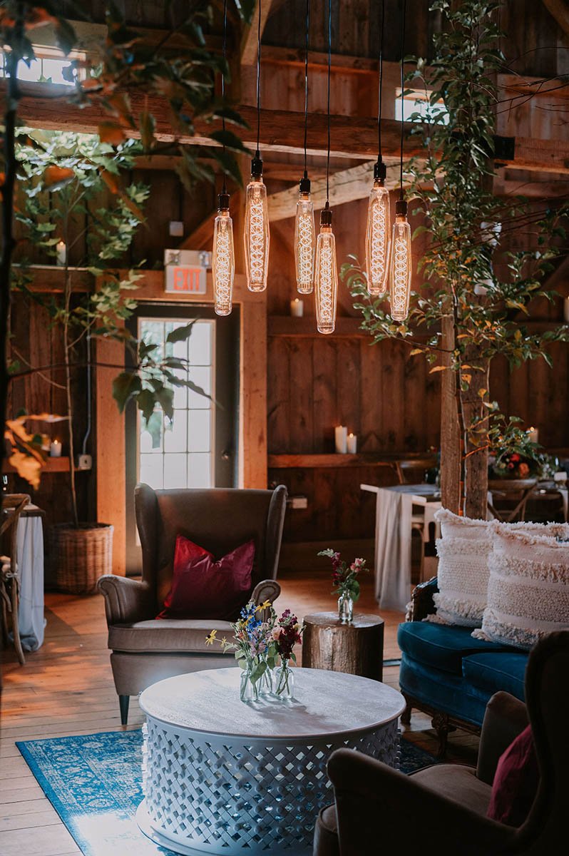 A cozy indoor setting with wooden walls and ceiling, featuring hanging vintage-style Edison bulbs, a black leather armchair with a red pillow, a round white coffee table with flowers, and various other chairs and plants.