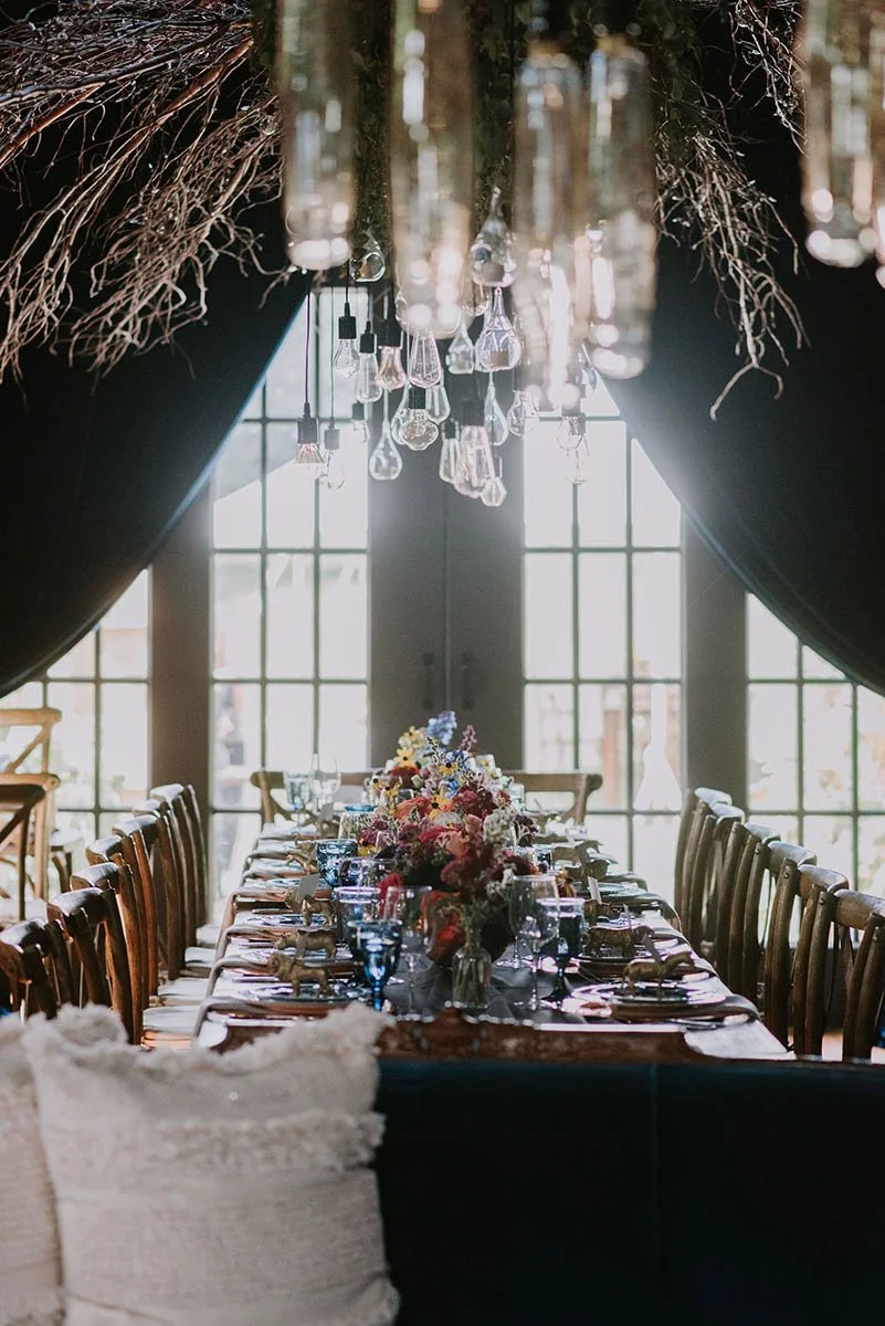 Long dining table set for a formal event with flower arrangements, glassware, and cutlery, in a room with large windows and hanging glass pendant lights.