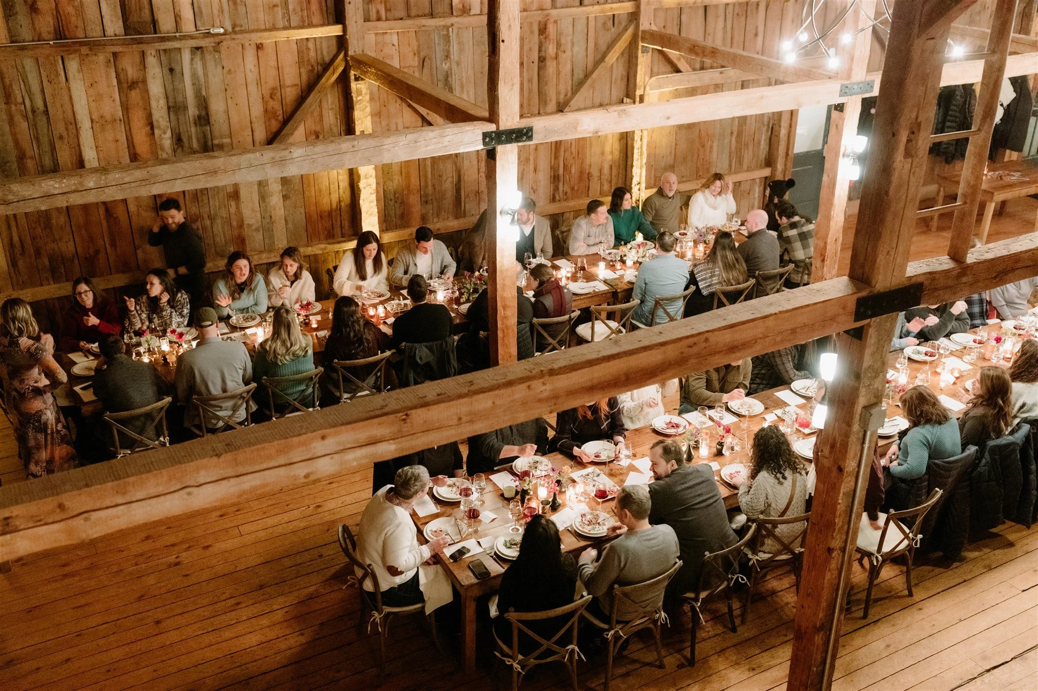 A group of people gathered around long wooden tables in a rustic, barn-like setting for a dinner or celebration, with warm lighting and decorated with flowers and candles.