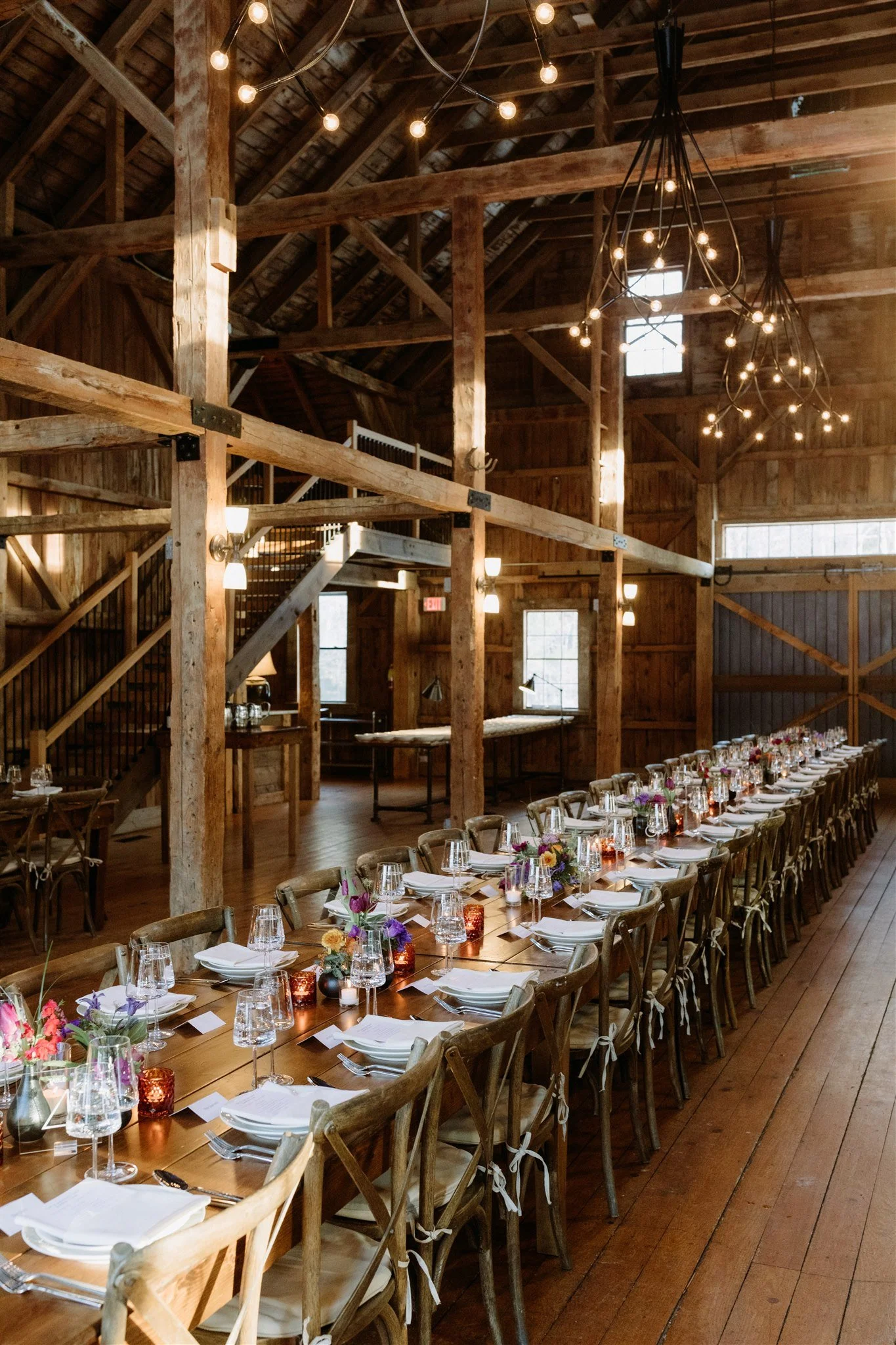 Long wooden banquet table set for a celebration in a rustic barn with string lights overhead and small floral arrangements.