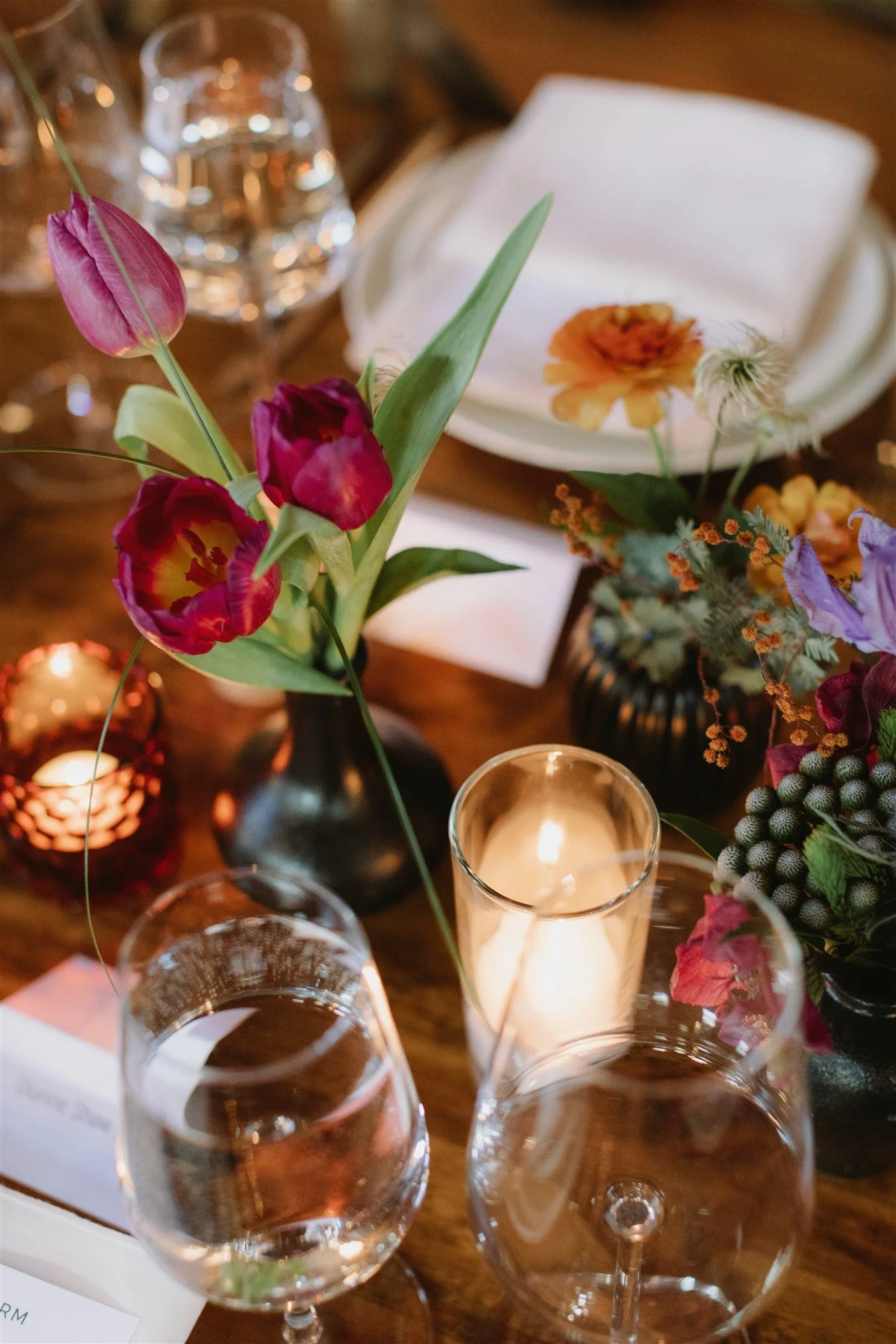 A table centerpiece featuring pink, purple, and yellow flowers in black vases, surrounded by wine glasses, a candle, and a white napkin on a wooden table.