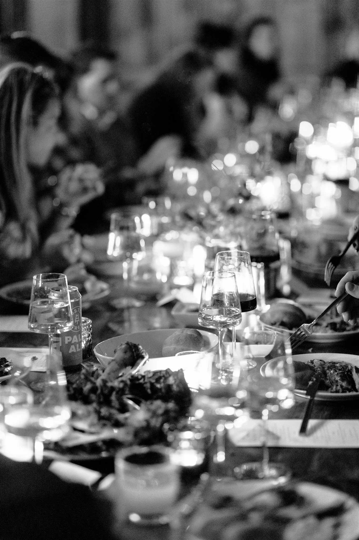 People dining at a long table with glasses and plates, in a dimly lit restaurant or gathering, in black-and-white photo.