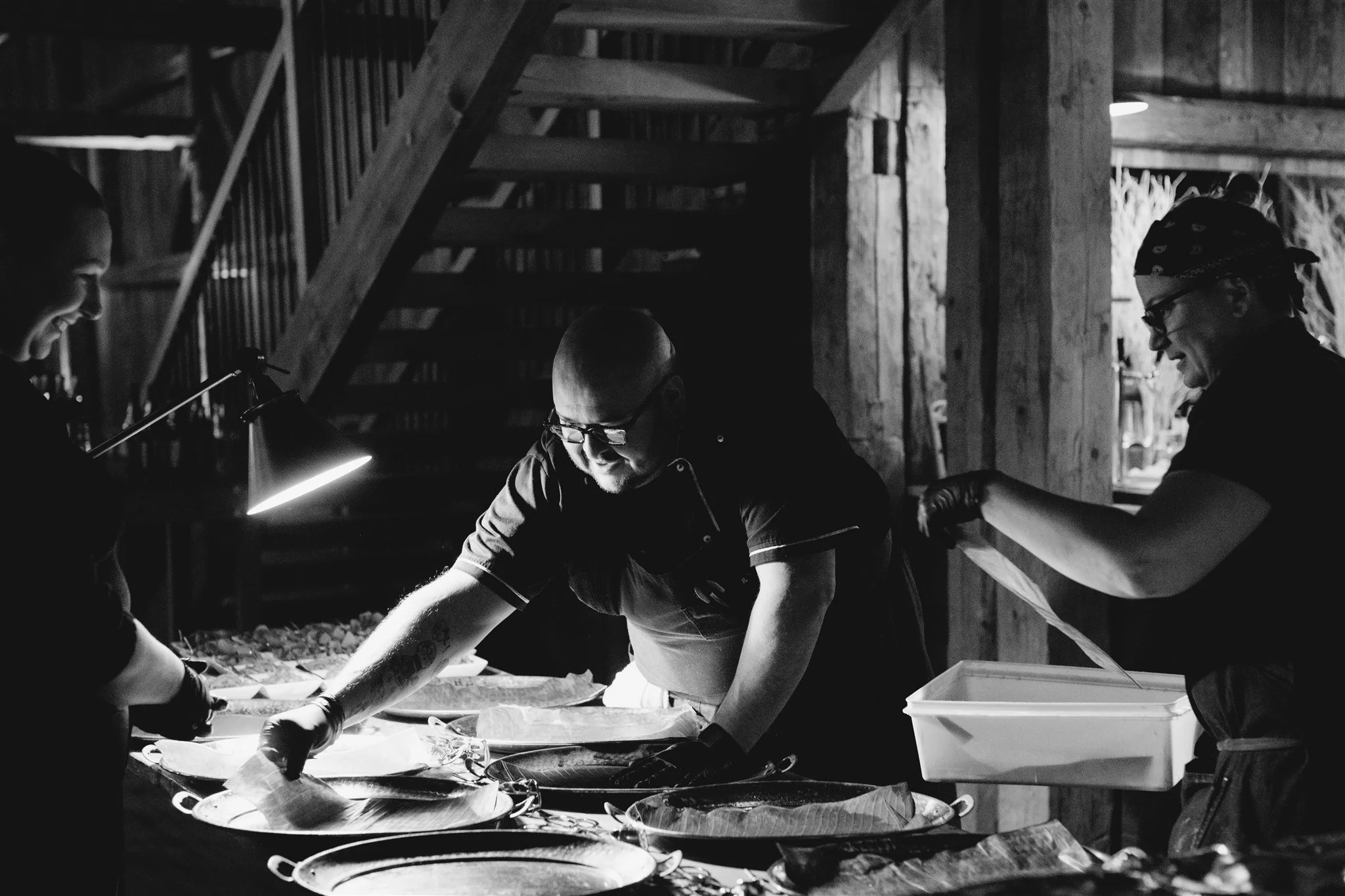 Three people working together in a dimly lit kitchen or food prep area, preparing food on a counter with trays and utensils, smiling and engaging with each other.