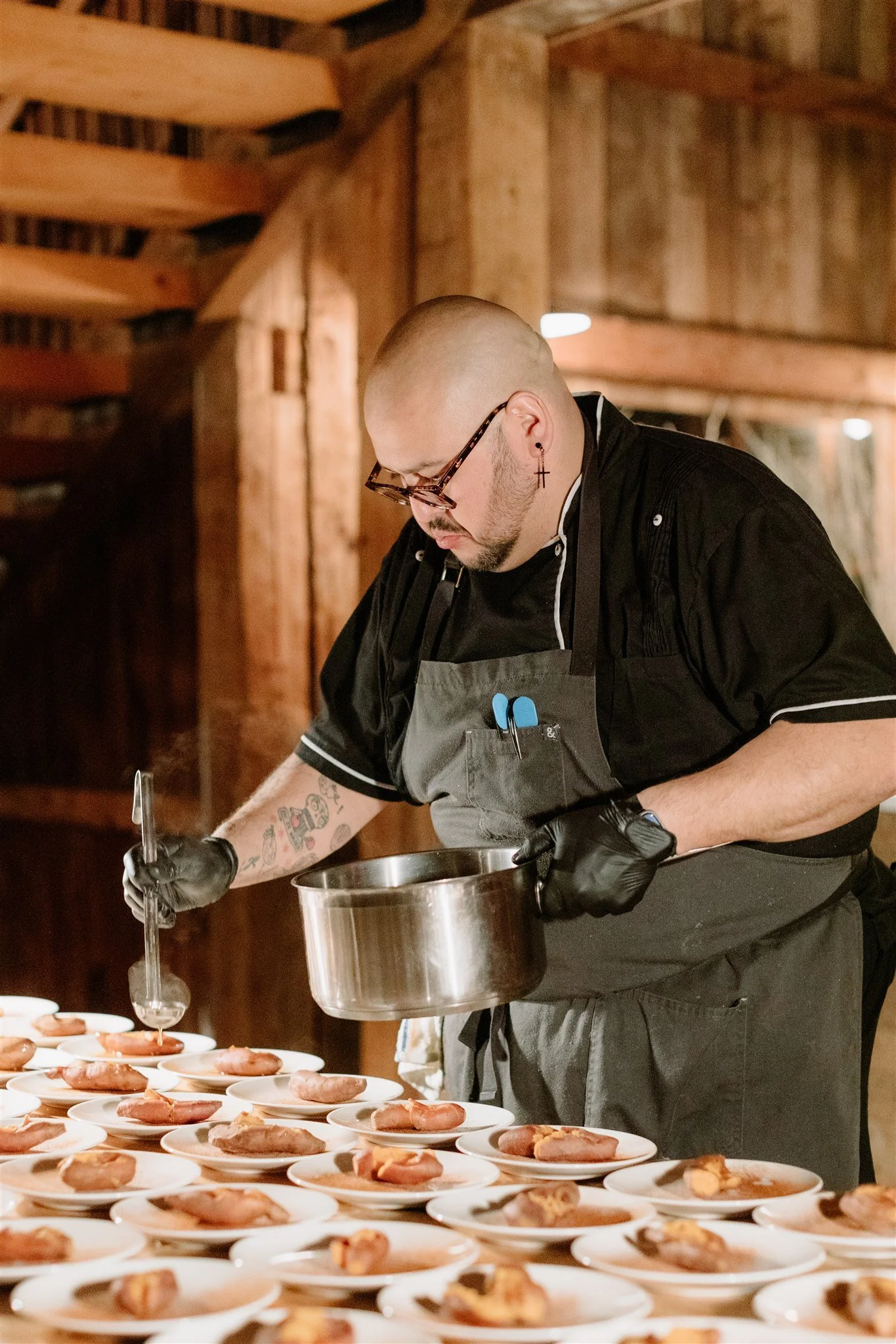 A chef in black clothing and glasses meticulously plates food onto small white dishes in a rustic wooden kitchen or restaurant setting.