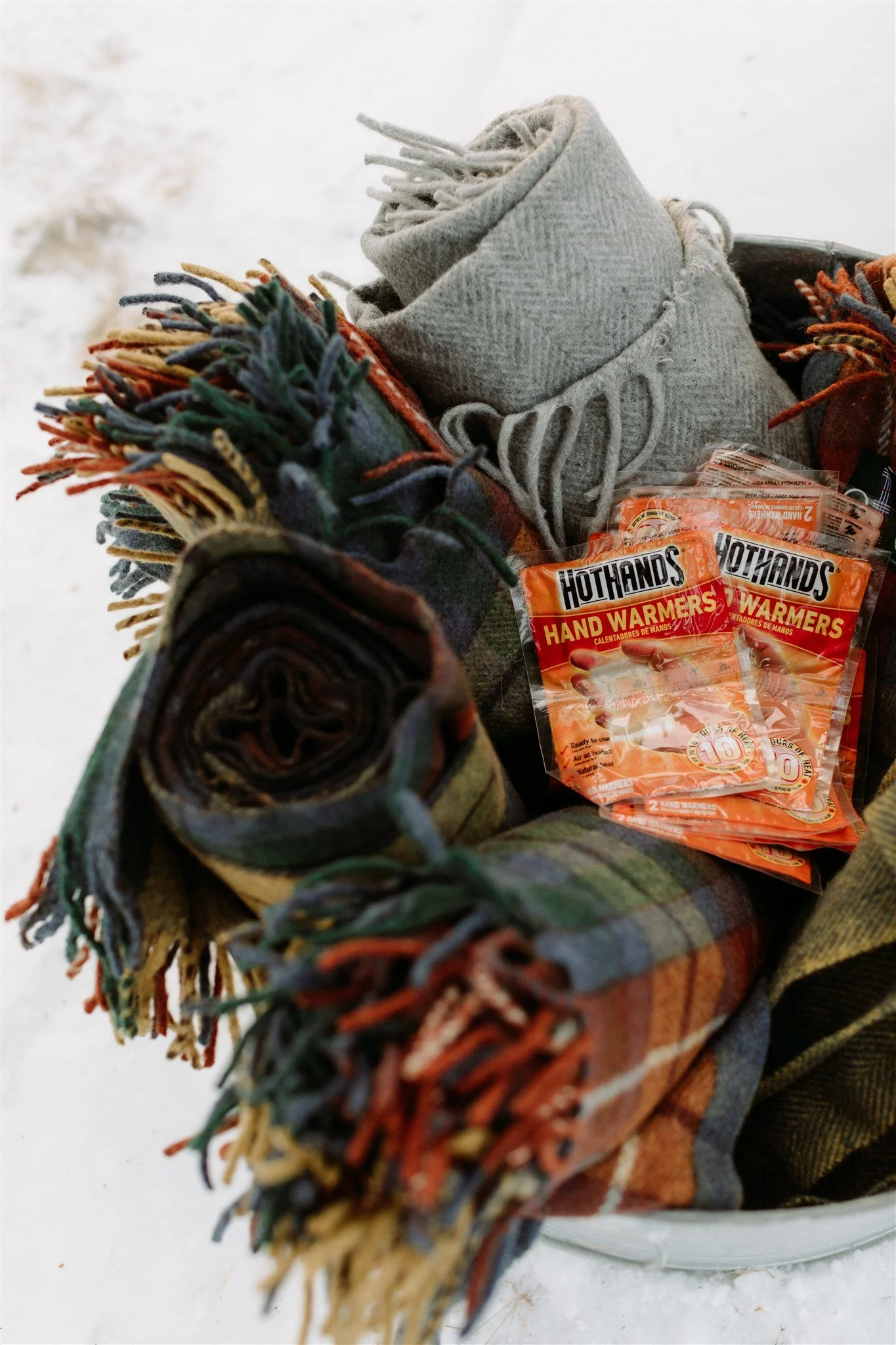 A collection of warm clothing including rolled-up scarves, a pair of gray knit socks, and multiple packets of hand warmers on a snowy surface.