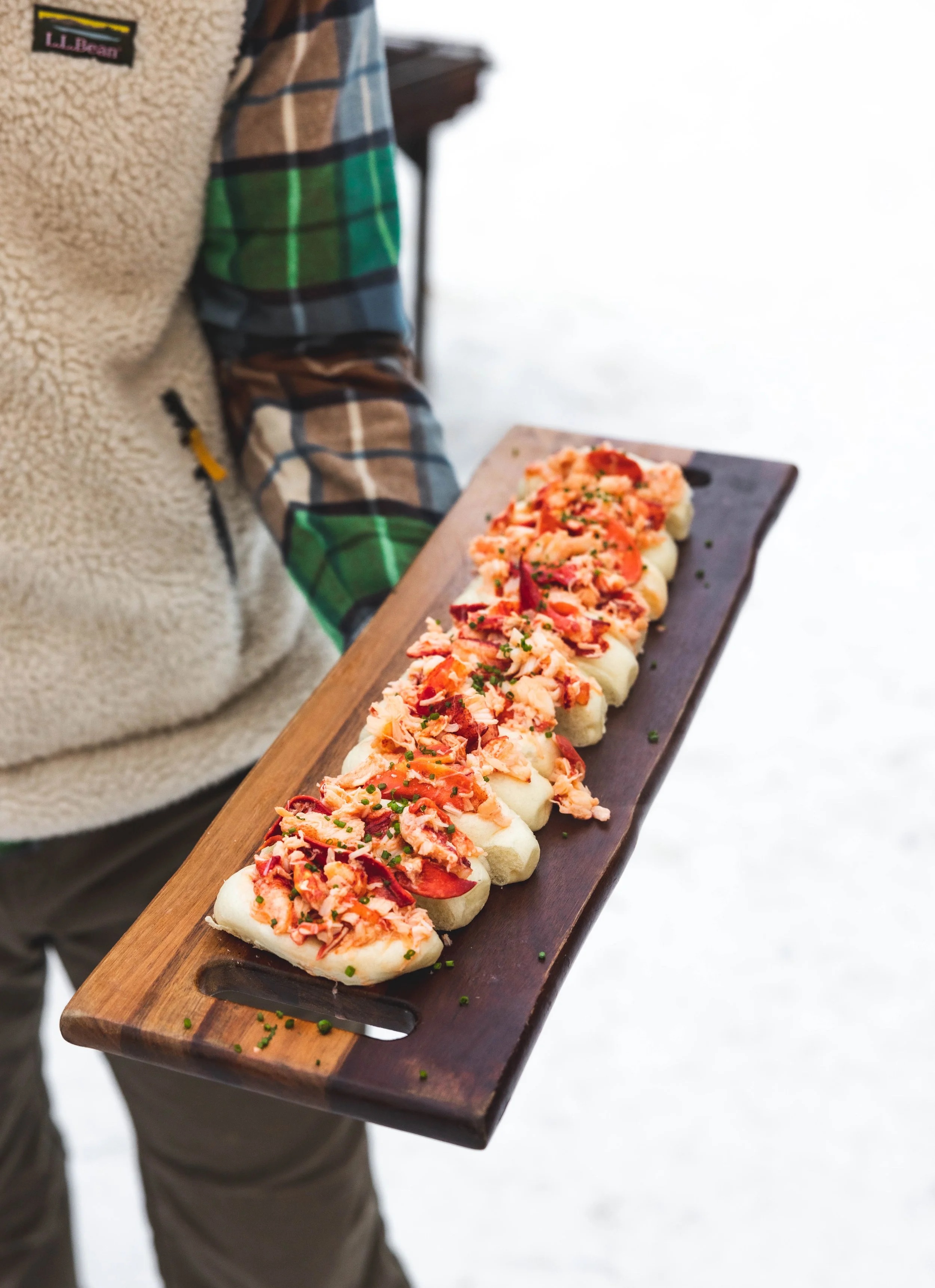 Person holding a wooden serving platter with small sandwiches topped with shredded seafood and herbs.