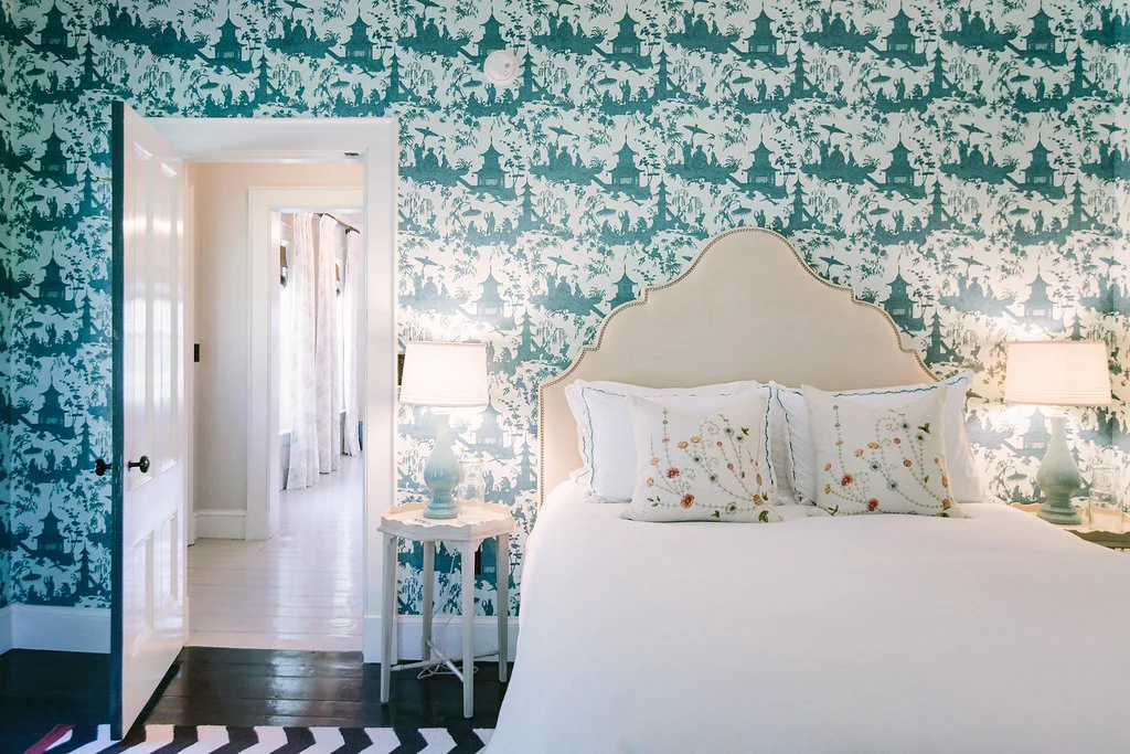 A bedroom with a bed, wicker headboard, white bedding, embroidered pillows, teal toile wallpaper, two bedside lamps, a small white side table, and doorway leading to a hallway with white curtains.