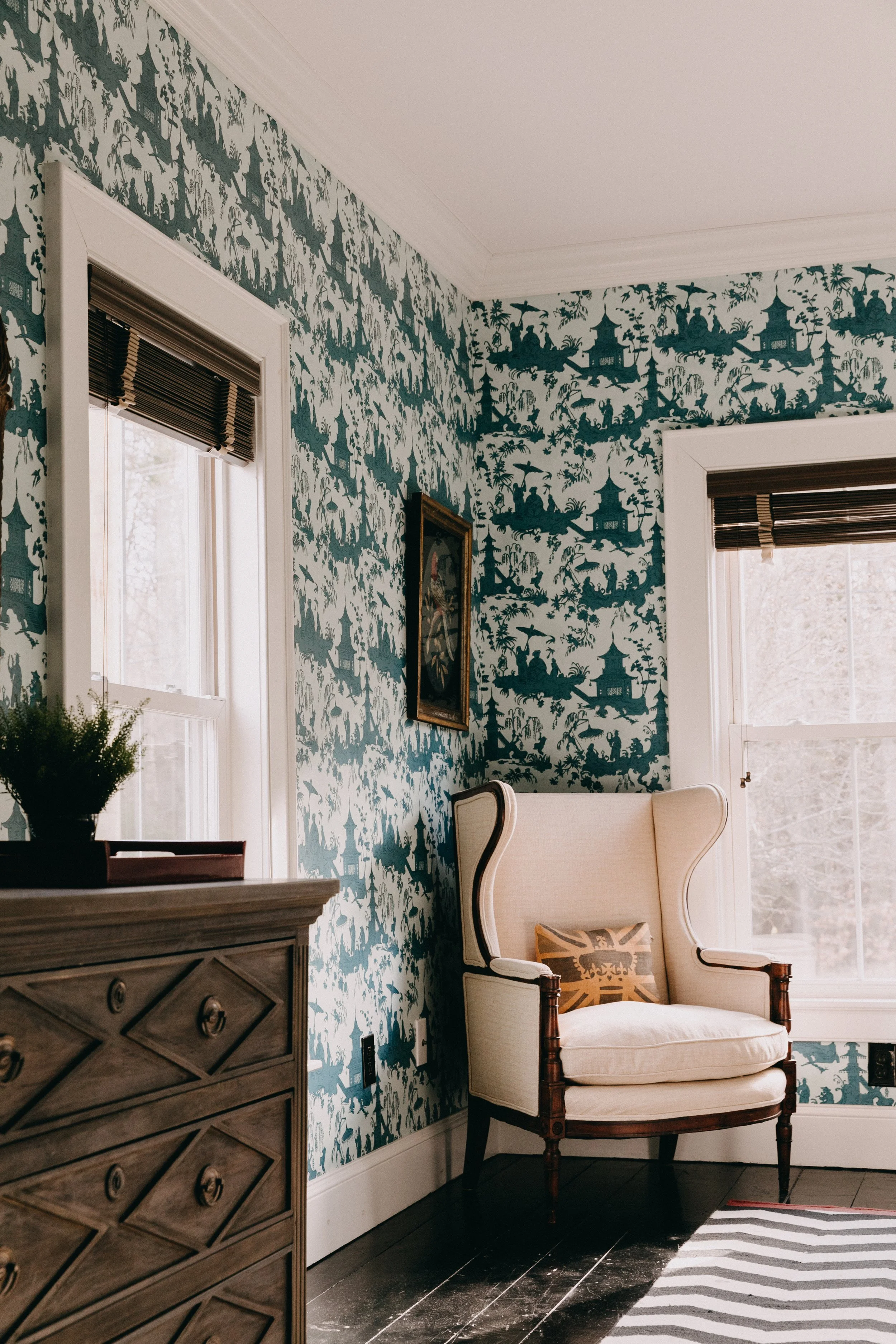 A cozy corner of a home with a white upholstered armchair with wooden trim, a decorative cushion, a wooden dresser with plants, and patterned wallpaper featuring a summer house scene with pagodas and trees.