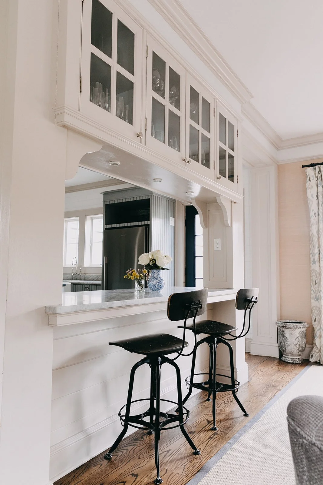 A kitchen view with a breakfast bar, two black bar stools, a vase with white flowers on the counter, and a view into the kitchen with a refrigerator and windows.