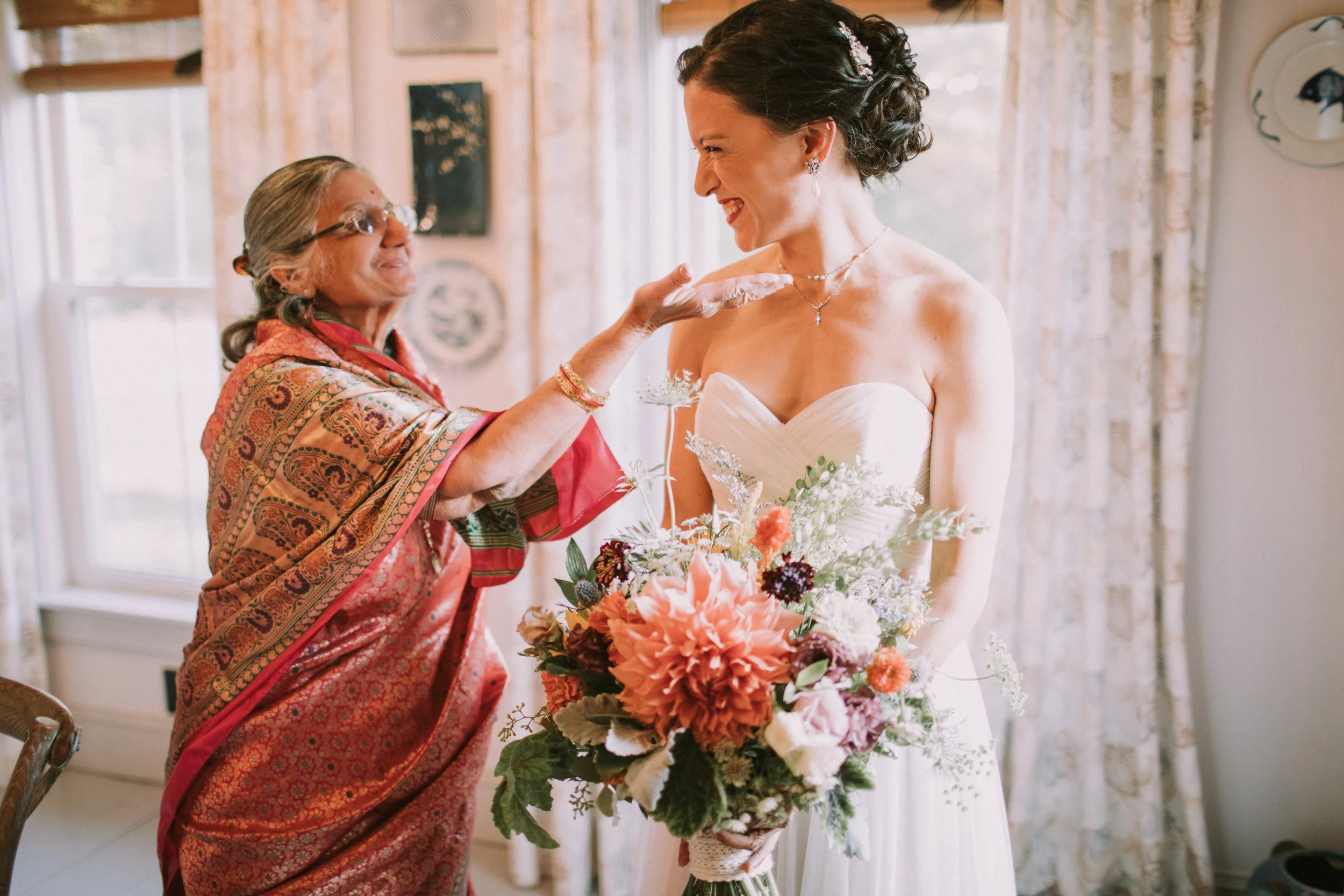 A bride with dark curly hair wearing a strapless white wedding dress, smiling, holding a large bouquet of pink, white, and green flowers, as an older woman with glasses and gray hair in a colorful sari touches her chin affectionately.